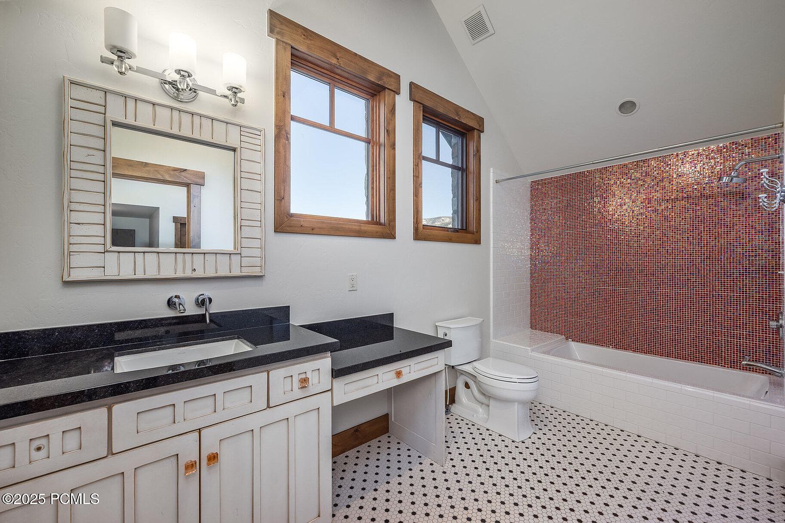 This is a well-lit bathroom featuring a vanity with a dark countertop and white cabinetry, a decorative mirror, and two windows with wooden frames. The bathroom also includes a toilet and a bathtub with a unique mosaic tile surround. The floor is tiled with a small black and white pattern, creating a classic and clean look.