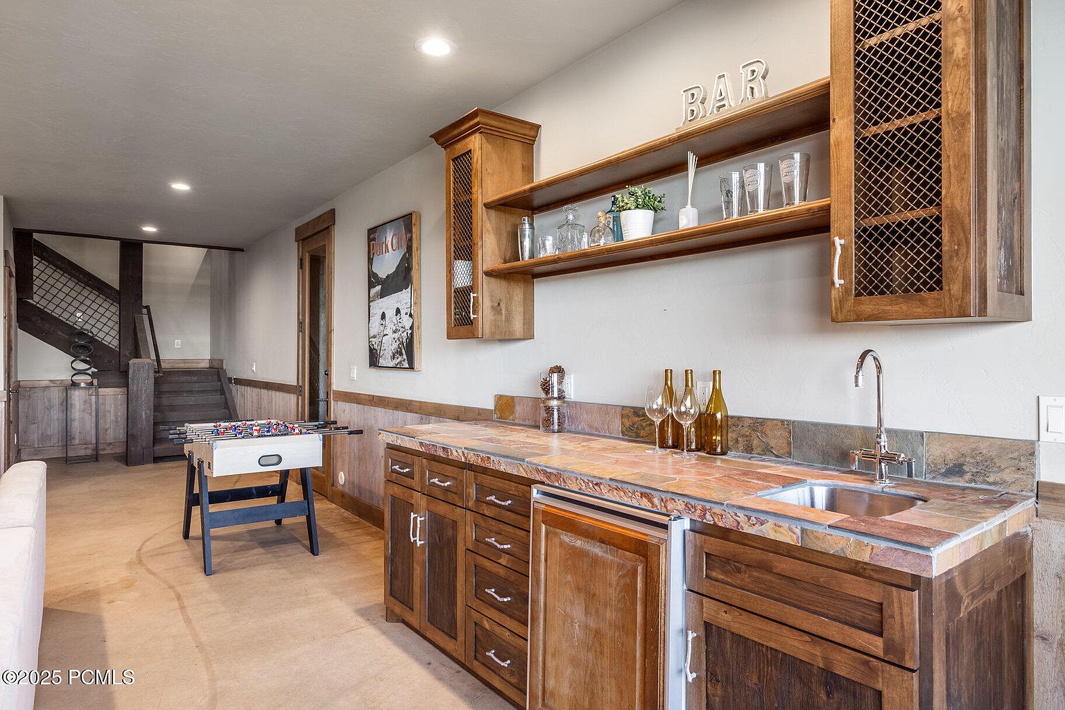 This interior shot showcases a basement area featuring a wet bar with wooden cabinetry and a unique tile countertop. The bar area includes a sink, wine storage, and shelving with glassware and decorative items. A foosball table adds a recreational element to the space, while the staircase in the background suggests access to other levels of the home.