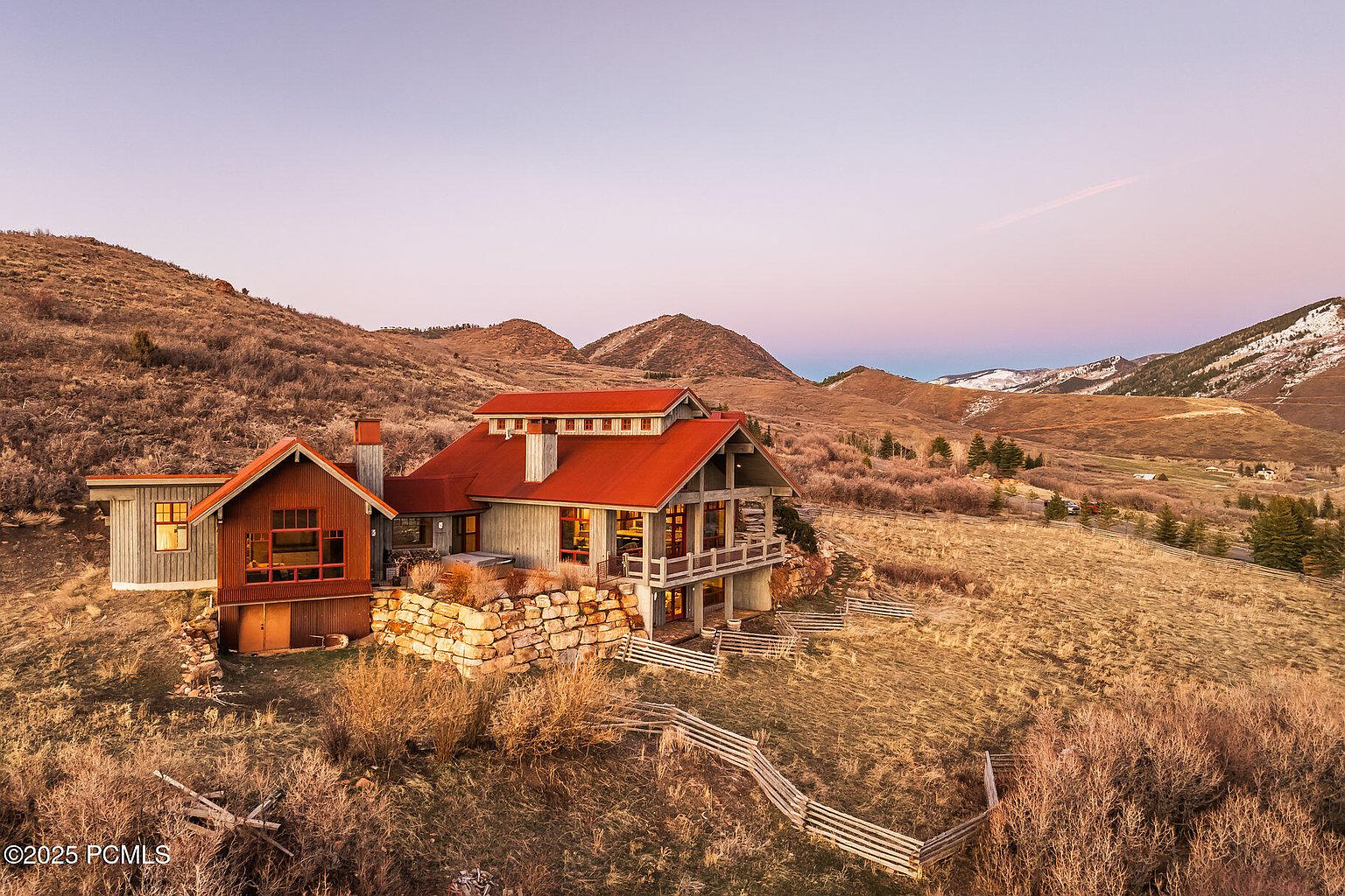 This aerial view showcases a stunning home nestled in a hilly, natural landscape. The house features a red roof, a combination of stone and wood exterior, and large windows that offer panoramic views. The surrounding terrain is a mix of dry grasses and shrubs, with mountains visible in the background, creating a sense of seclusion and natural beauty.