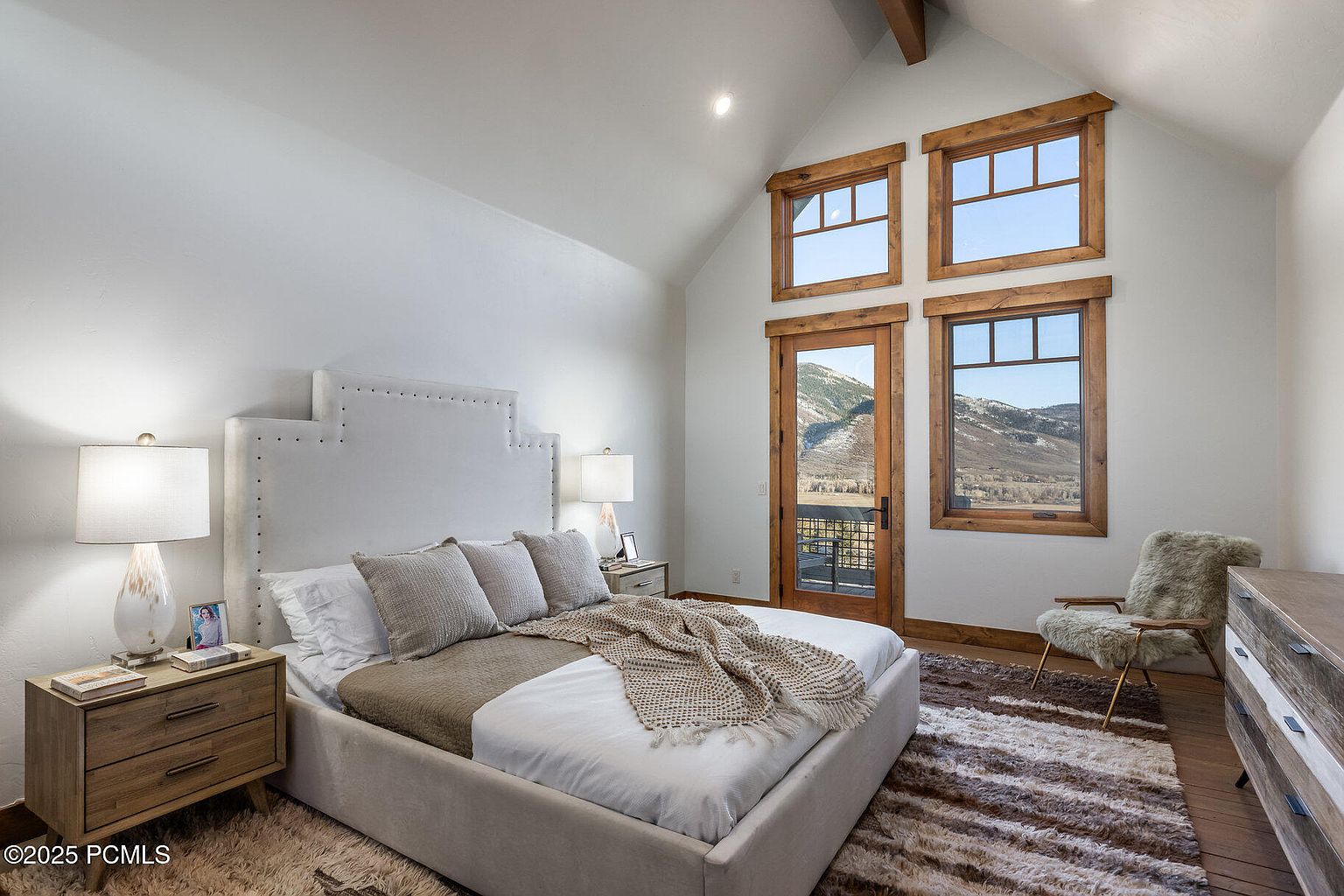 This is a well-lit primary bedroom featuring a large bed with a light-colored upholstered headboard and neutral bedding. The room has a high, vaulted ceiling with exposed beams and a set of windows and a door that offer a view of the mountains. The room is decorated in a neutral color palette with wooden accents, creating a cozy and inviting atmosphere.