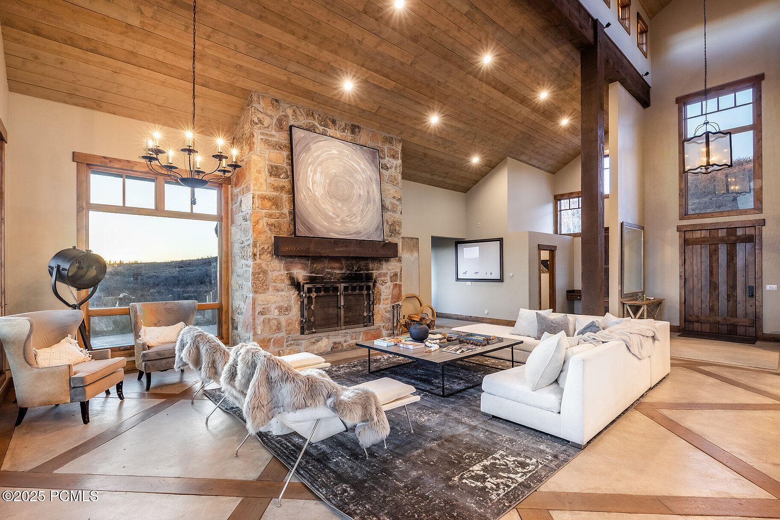 This is a spacious living room featuring a stone fireplace with a large art piece above it, a white sectional sofa, and two accent chairs. The room has a high, wood-paneled ceiling with recessed lighting and large windows that offer natural light. The flooring is a combination of concrete and wood, adding to the rustic yet modern aesthetic.