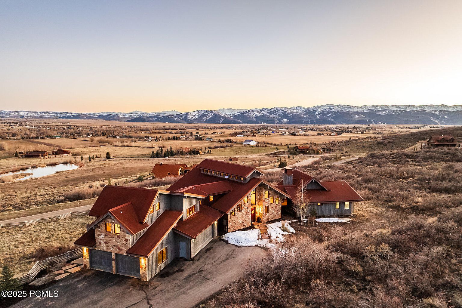 This aerial view showcases a sprawling estate nestled in a valley with mountains in the background. The home features a combination of stone and wood siding, with a red roof and multiple gables. The property includes a long driveway, some snow patches, and natural landscaping, creating a sense of privacy and seclusion.