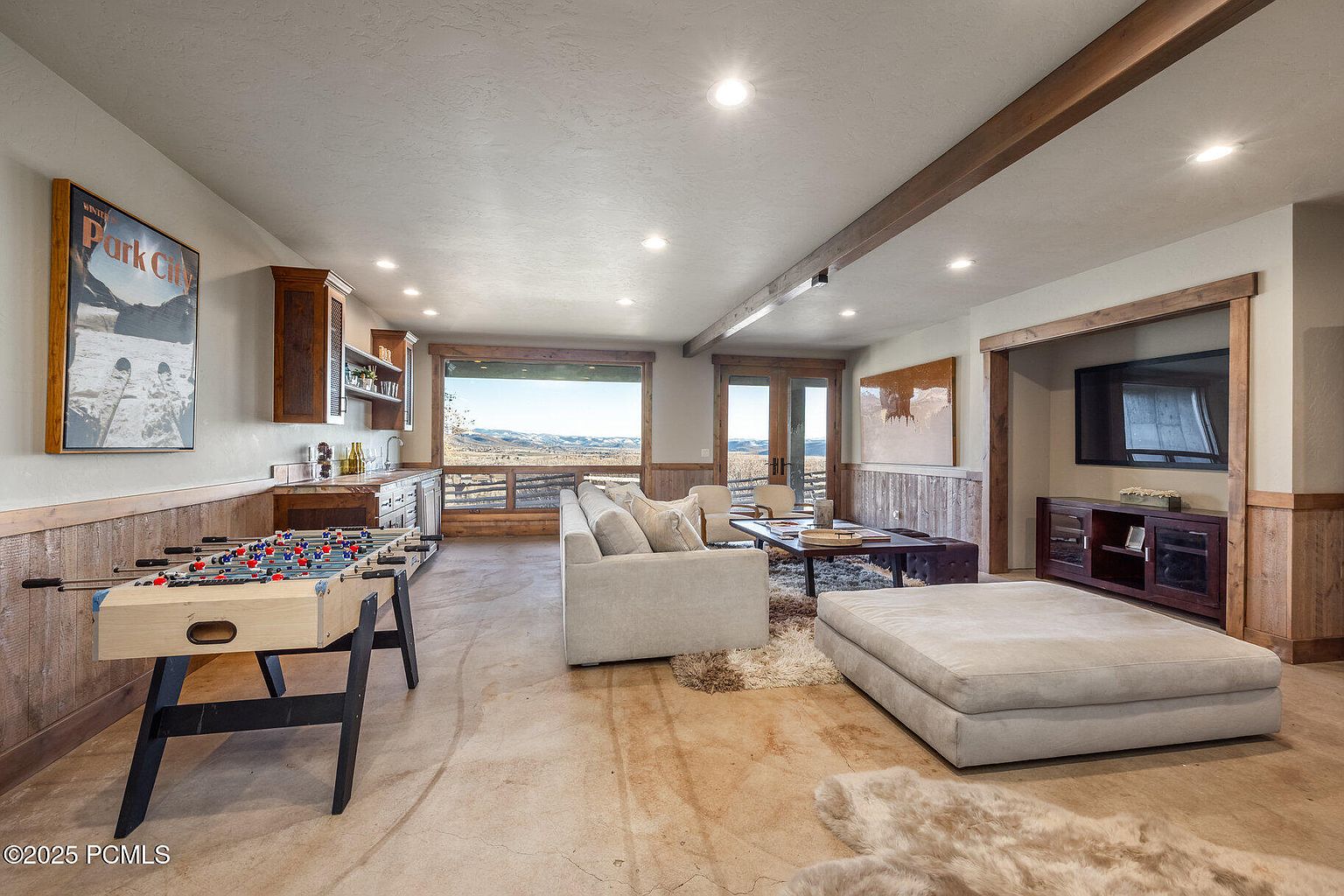 This interior shot showcases a spacious living room with a rustic-modern aesthetic. The room features a foosball table, a wet bar, a comfortable seating area with a large ottoman, and a media center with a television. Wood accents and trim add warmth, while large windows offer scenic views, creating an inviting and functional space.
