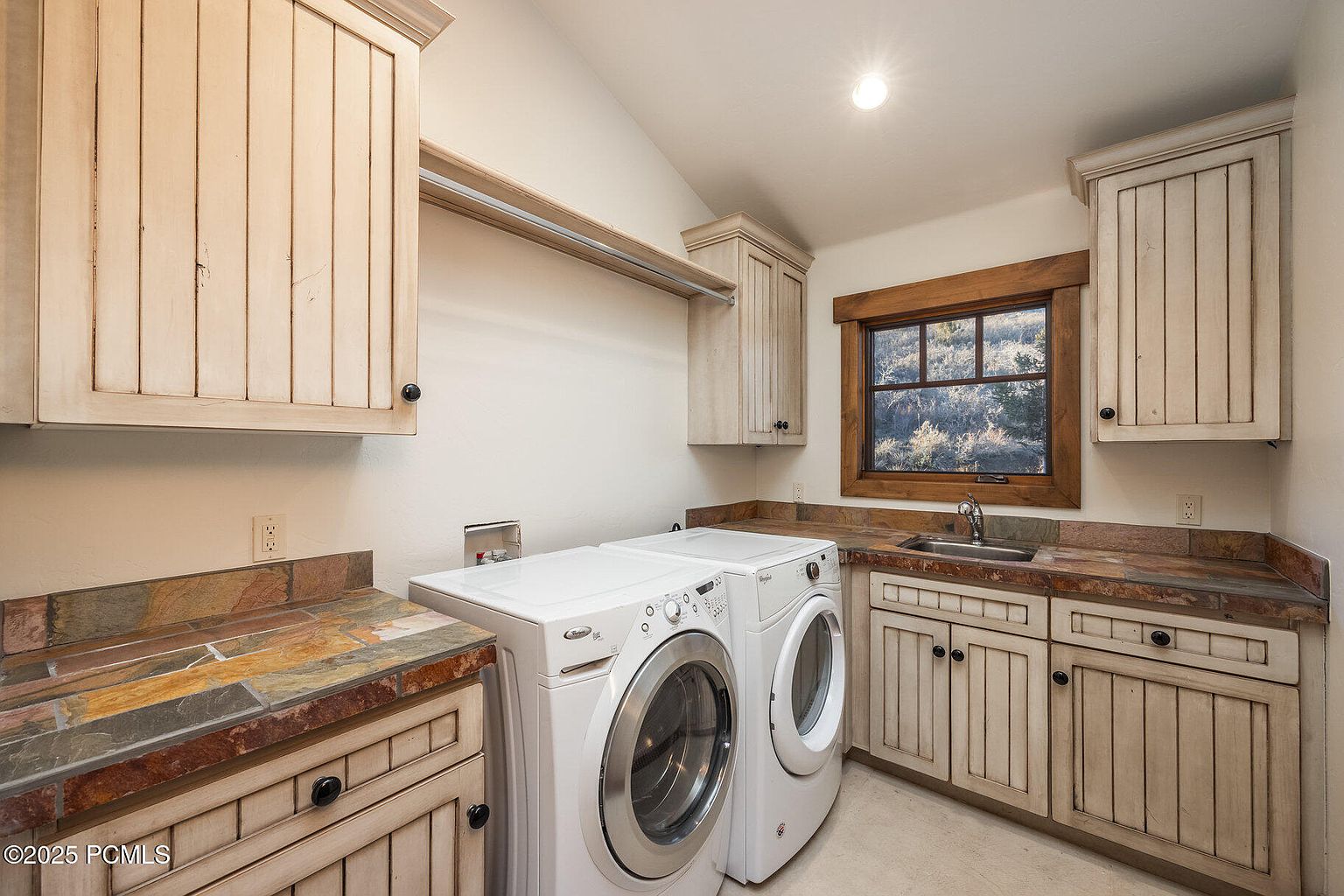 This is a well-organized laundry room featuring a front-loading washer and dryer set, custom cabinetry with a distressed finish, and a countertop with a unique stone pattern. A window provides natural light, and a hanging rod offers space for drying clothes. The room has a functional yet stylish design, perfect for a modern home.