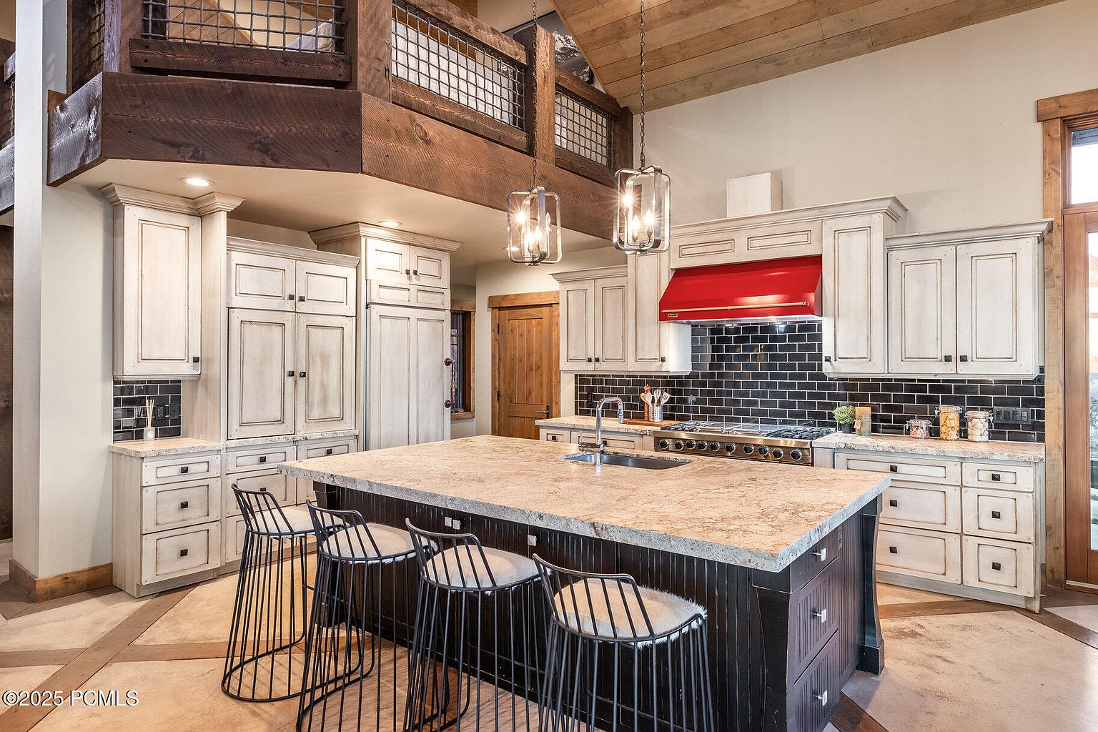 This is a well-lit kitchen featuring a large island with seating, custom cabinetry with a distressed finish, and a contrasting dark backsplash. The kitchen also includes a red range hood and modern pendant lighting. The overall impression is a blend of rustic charm and modern elegance.
