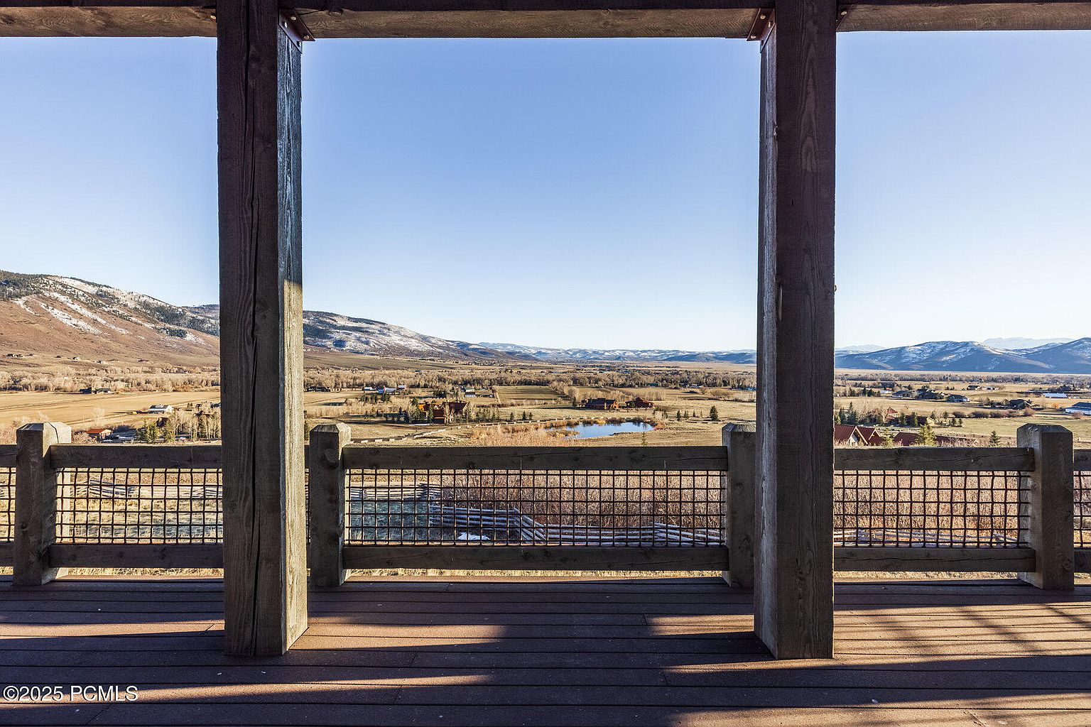 This image showcases a scenic view from a wooden deck or balcony, framed by sturdy wooden posts and railings. The landscape features a valley with scattered houses, a small pond, and distant mountains under a clear blue sky. The deck itself is made of composite wood, suggesting a durable and low-maintenance outdoor space.
