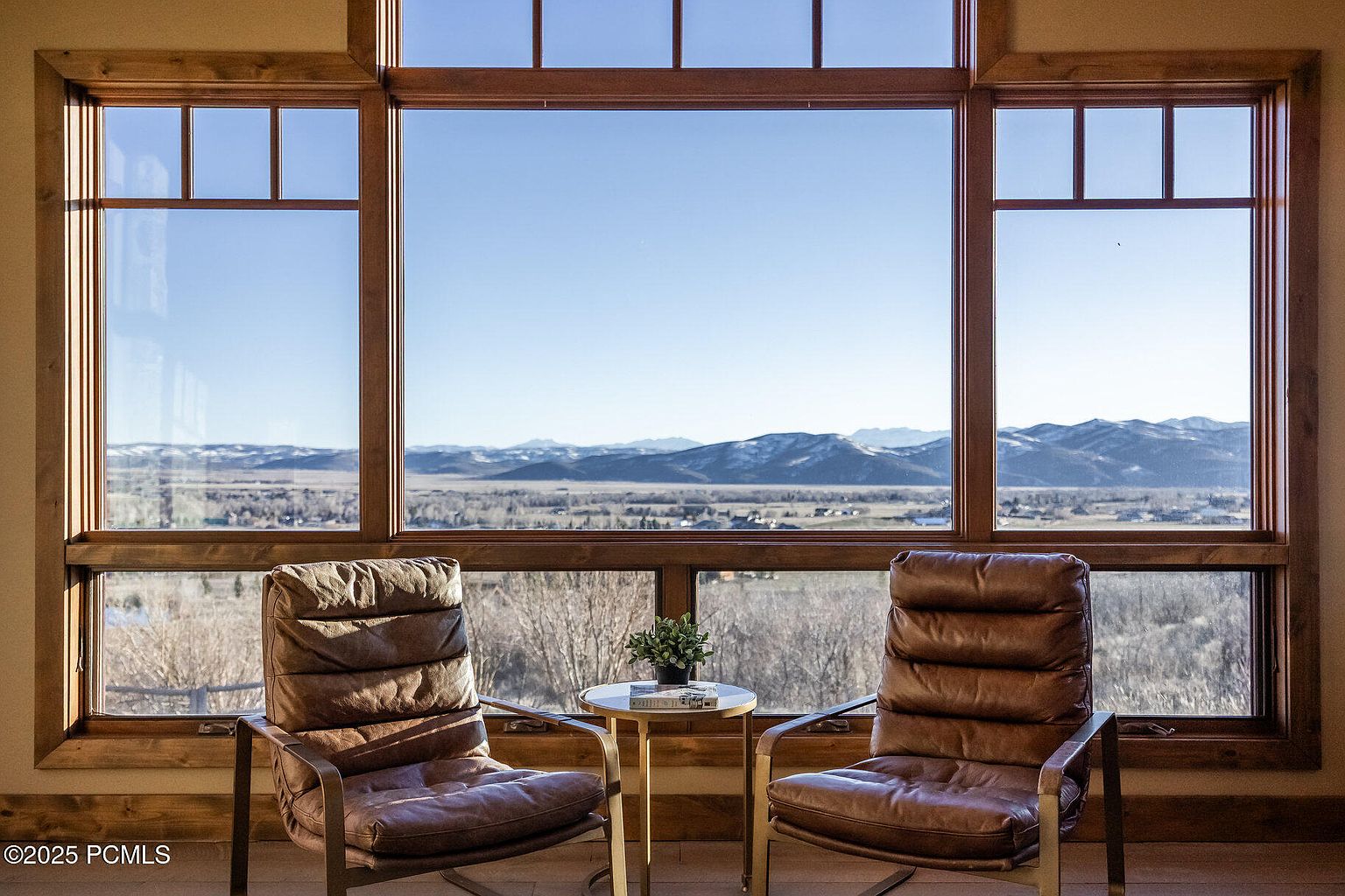 This interior shot showcases a cozy living room setup with two leather armchairs flanking a small side table, all positioned in front of a large window offering a scenic view of distant mountains and a valley. The natural wood window frame adds warmth, while the chairs invite relaxation and contemplation of the landscape, creating an inviting and serene atmosphere.