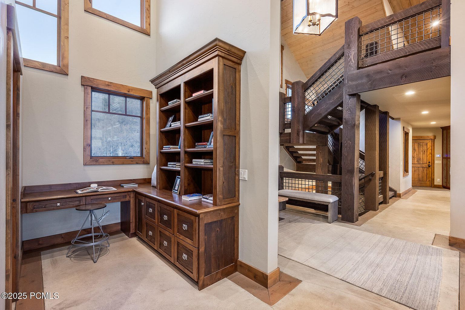 This interior shot showcases a cozy office/study area with custom wooden built-ins, including a desk and shelving unit. The room features natural light from a window, complemented by a rustic staircase in the background, adding character to the space. The overall impression is one of warmth and functionality, ideal for a home office.