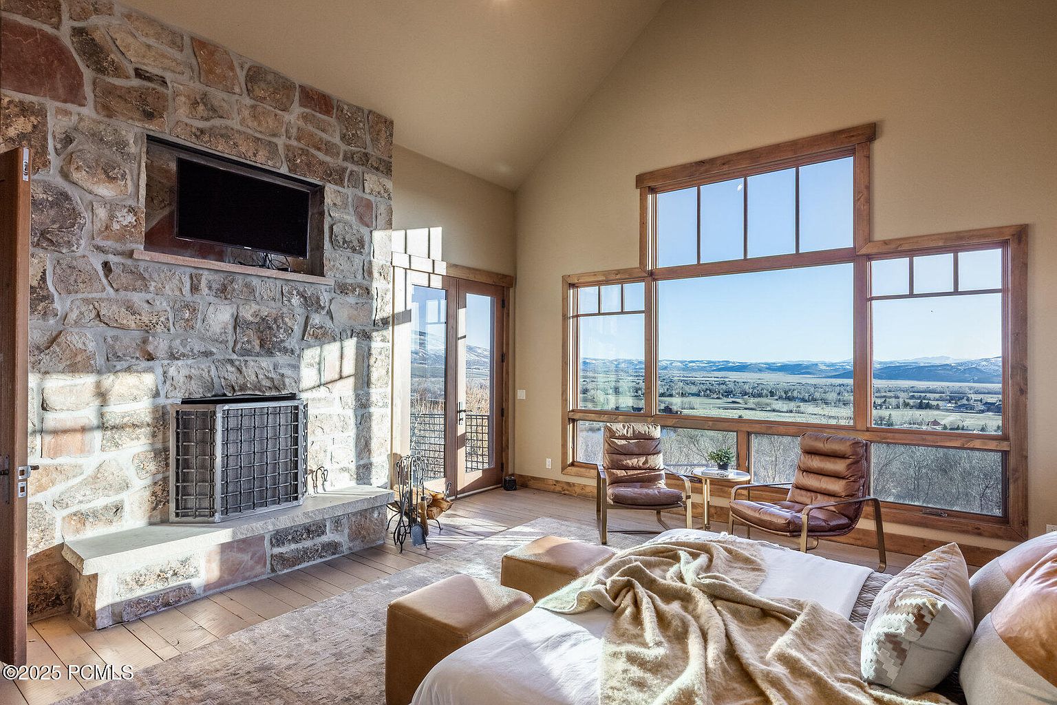 This is a primary bedroom featuring a stone fireplace with a mounted television, large windows with a scenic view, and comfortable seating. The room is well-lit with natural light, creating a warm and inviting atmosphere. The bed is neatly made with a throw blanket and decorative pillows.