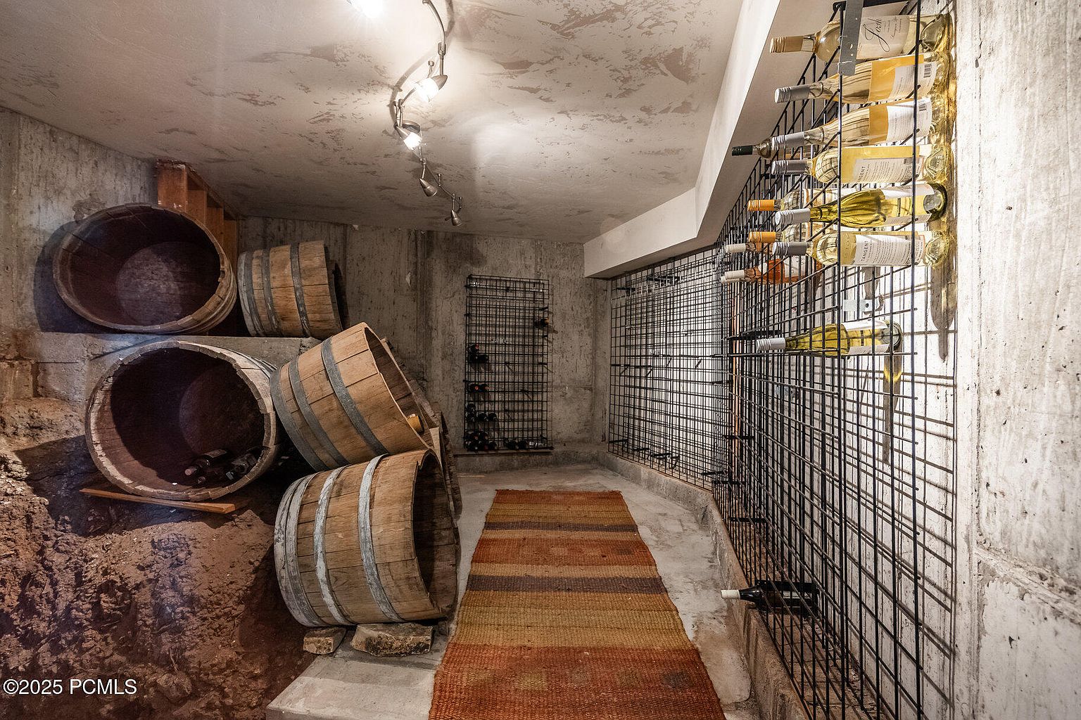 This is a wine cellar featuring a rustic design with exposed concrete walls and a collection of wine barrels. A metal grid wine rack displays various bottles, and a textured rug adds warmth to the space. The lighting is soft, creating an intimate and inviting atmosphere for wine storage and tasting.