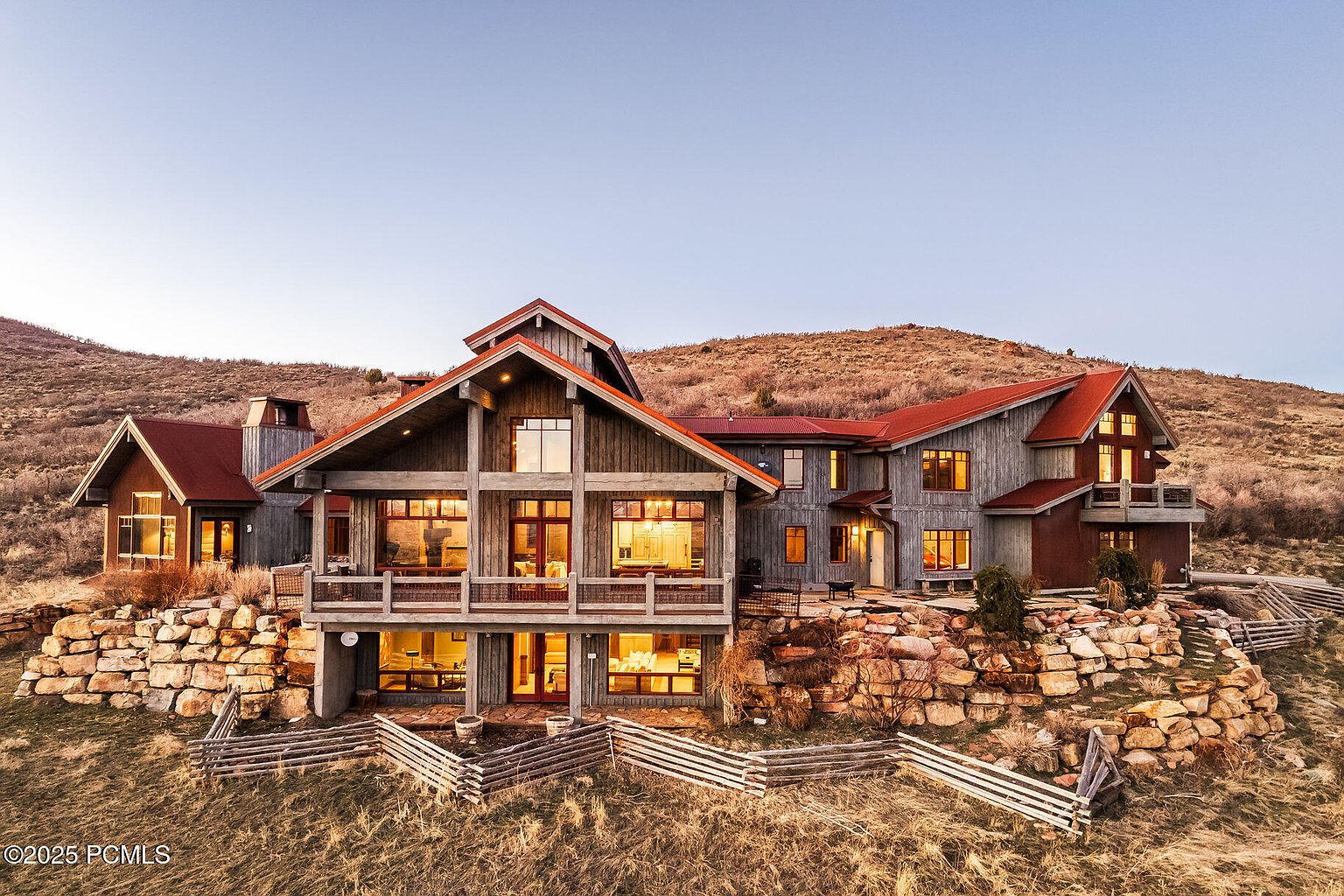 This is a front exterior view of a large, multi-level mountain home. The house features a combination of wood and stone elements, with a red roof and numerous windows that provide ample natural light. The property is situated on a hillside, with a rustic wooden fence and natural rock landscaping adding to its charm.