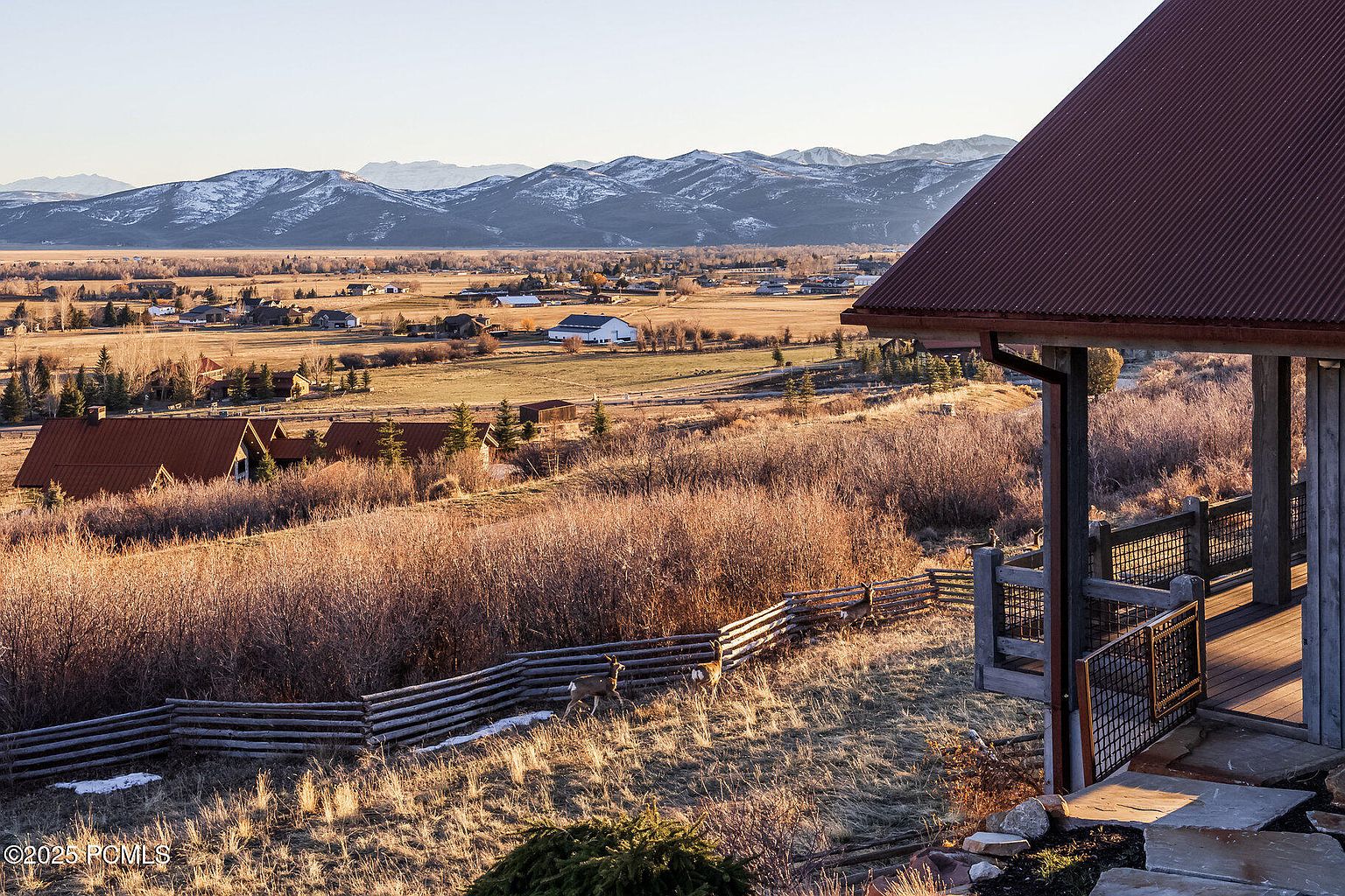 This image showcases a scenic view from a patio or deck, featuring a rustic wooden fence and a glimpse of deer in the foreground. In the distance, a valley with scattered houses and snow-capped mountains creates a picturesque backdrop. The overall impression is one of tranquility and natural beauty, highlighting the property's connection to its surroundings.