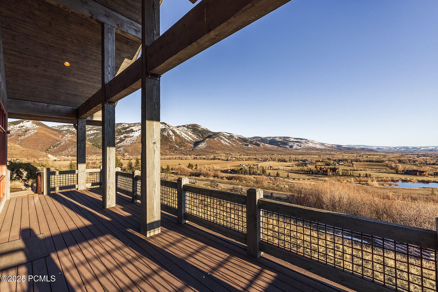 This image showcases a spacious deck or balcony with wooden flooring and a rustic railing, offering panoramic views of a valley and distant mountains. The structure features sturdy wooden beams and posts, creating a sense of natural elegance. The clear blue sky enhances the serene and inviting atmosphere, making it an ideal outdoor living space.