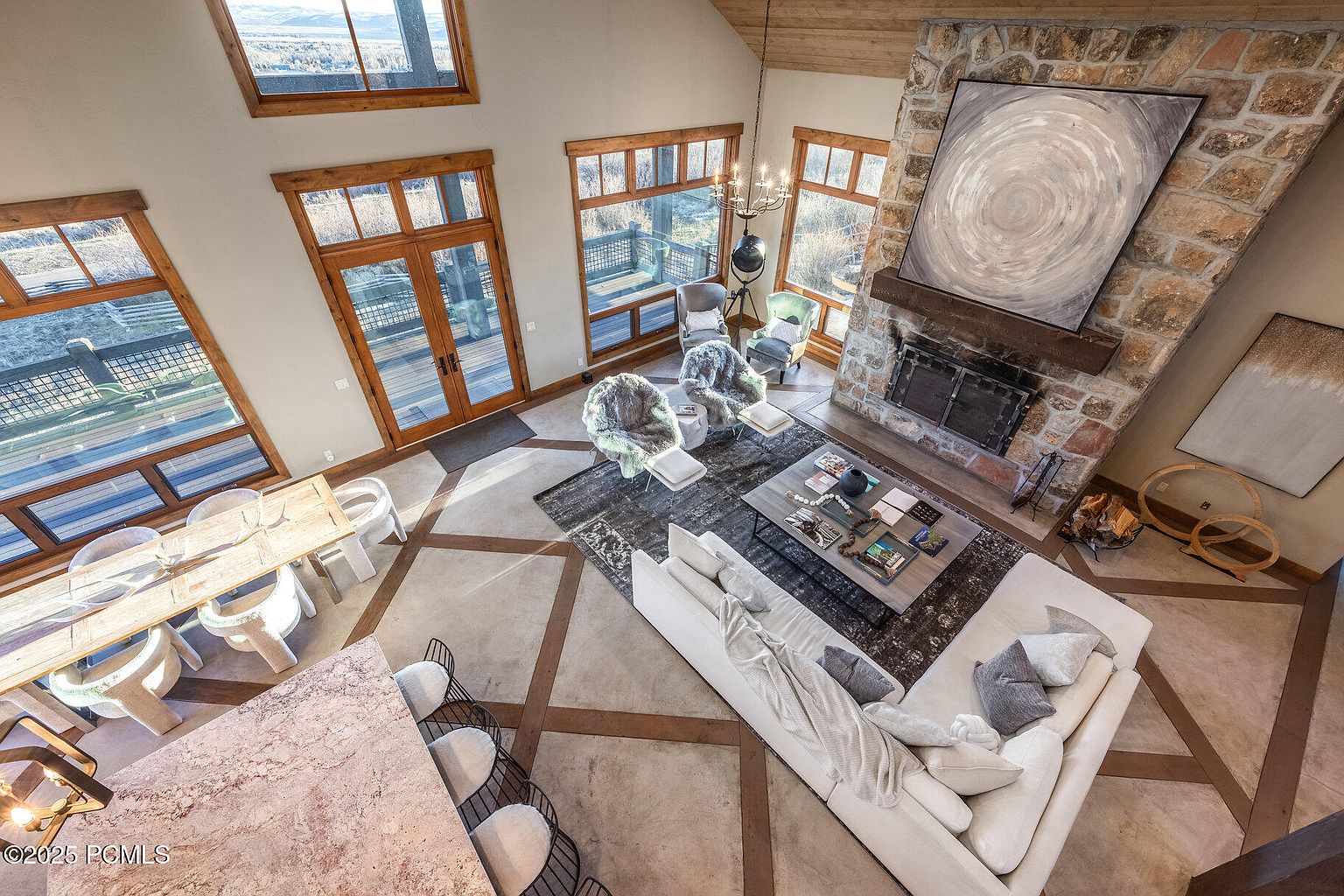 This is an elevated view of a luxurious living room featuring a stone fireplace with a large abstract painting above it. The room is well-lit with natural light streaming through large windows, highlighting the hardwood floors and a comfortable white sectional sofa. The space is designed with a blend of rustic and modern elements, creating a warm and inviting atmosphere.