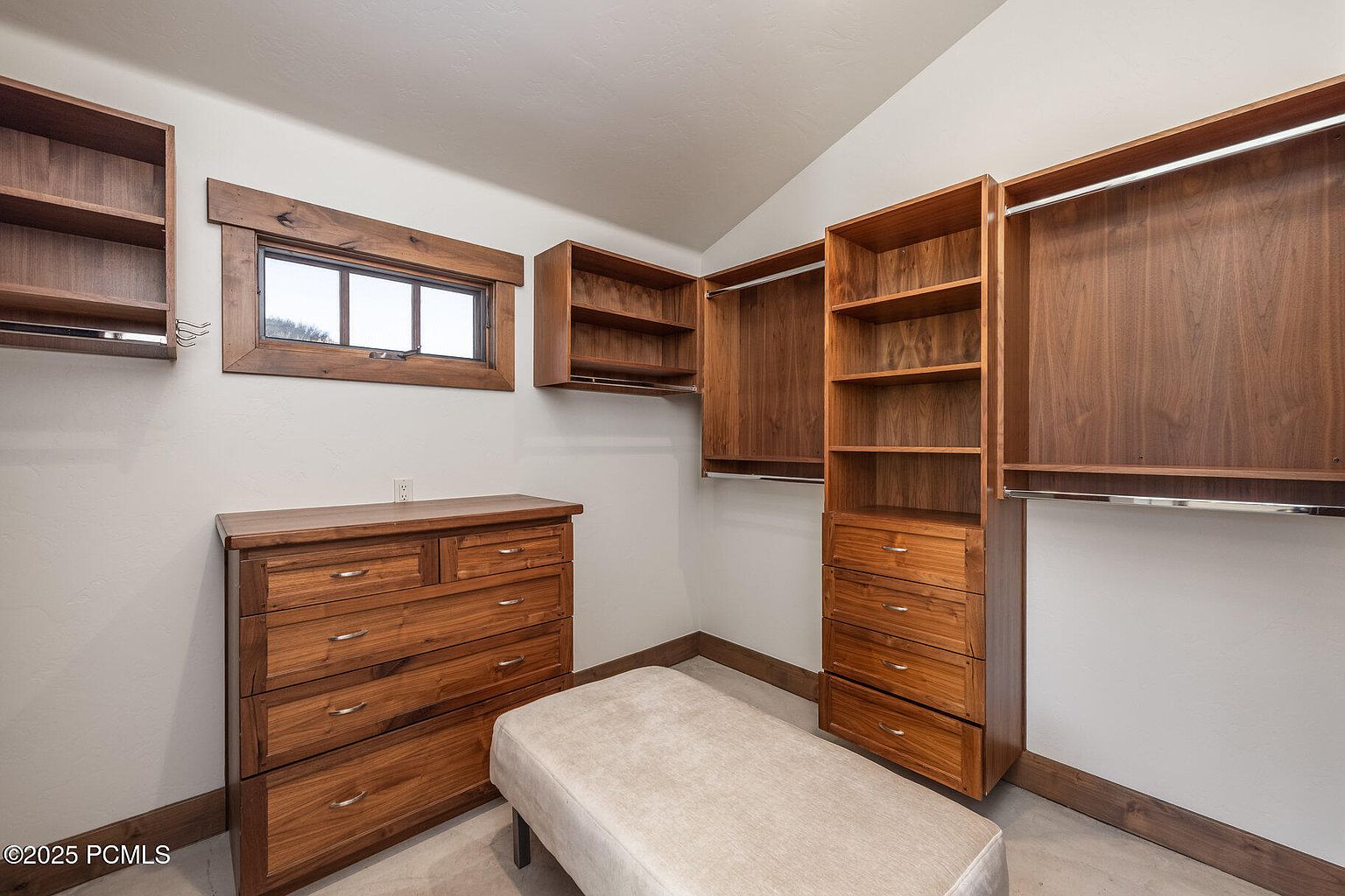 This is a well-organized walk-in closet featuring custom wooden cabinetry and shelving. A dresser and a cushioned bench provide additional storage and seating. The space is illuminated by natural light from a window, creating a warm and inviting atmosphere.