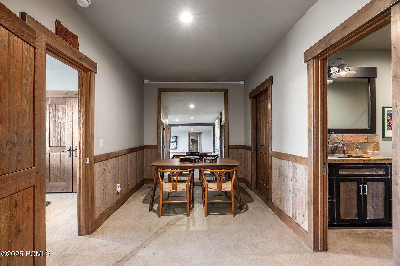 This interior shot showcases a hallway with rustic wood trim and wainscoting. A wooden table with chairs sits in the center, reflecting in a large mirror at the end of the hall. Doorways on either side lead to other rooms, one revealing a glimpse of a kitchenette area.