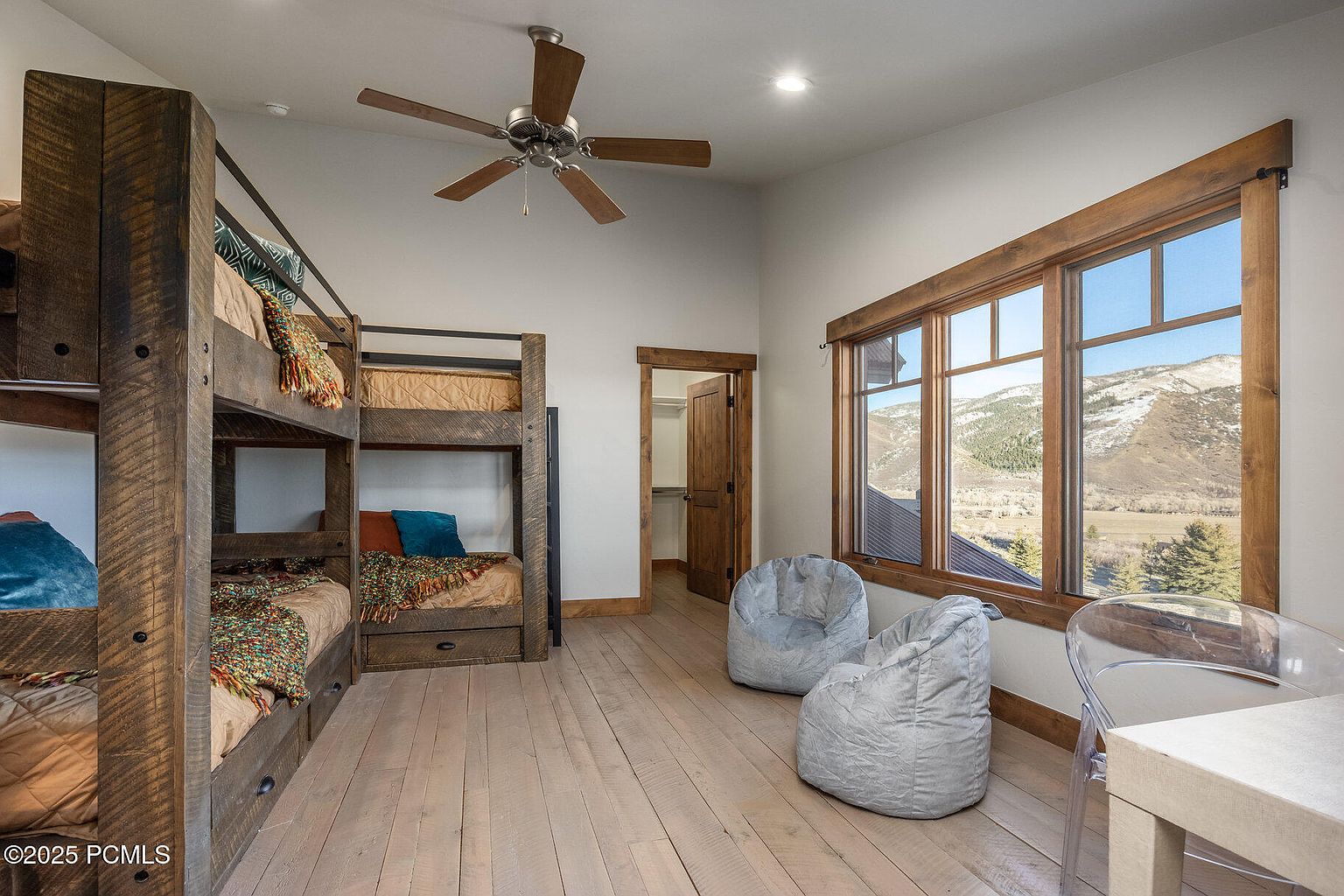 This is a cozy guest bedroom featuring custom-built wooden bunk beds, providing ample sleeping space. The room is well-lit with natural light streaming through a large window that offers scenic mountain views. Two beanbag chairs and a small desk create a comfortable and inviting atmosphere, complemented by the light wood flooring.