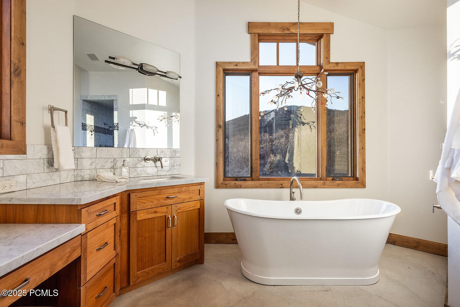 This is a well-lit primary bathroom featuring a freestanding white bathtub positioned in front of a large window with a wooden frame, offering a view of the outdoors. The bathroom also includes a wooden vanity with a marble countertop and a large mirror. The overall style is luxurious and inviting, with a focus on natural light and high-end finishes.
