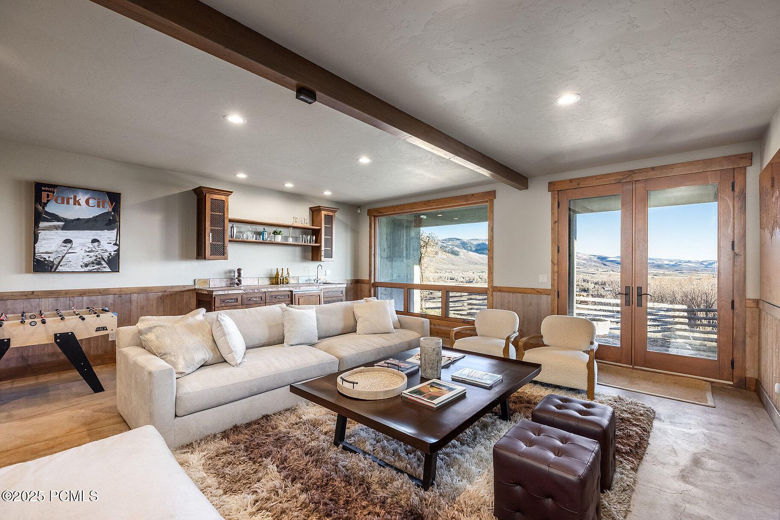 This inviting living room features a comfortable beige sectional sofa, a dark wood coffee table with decorative items, and two armchairs facing a large window with scenic views. The room is accented by a textured rug, wooden beams, and a built-in bar area, creating a cozy and luxurious atmosphere perfect for relaxation and entertaining. The presence of a foosball table adds a playful touch.