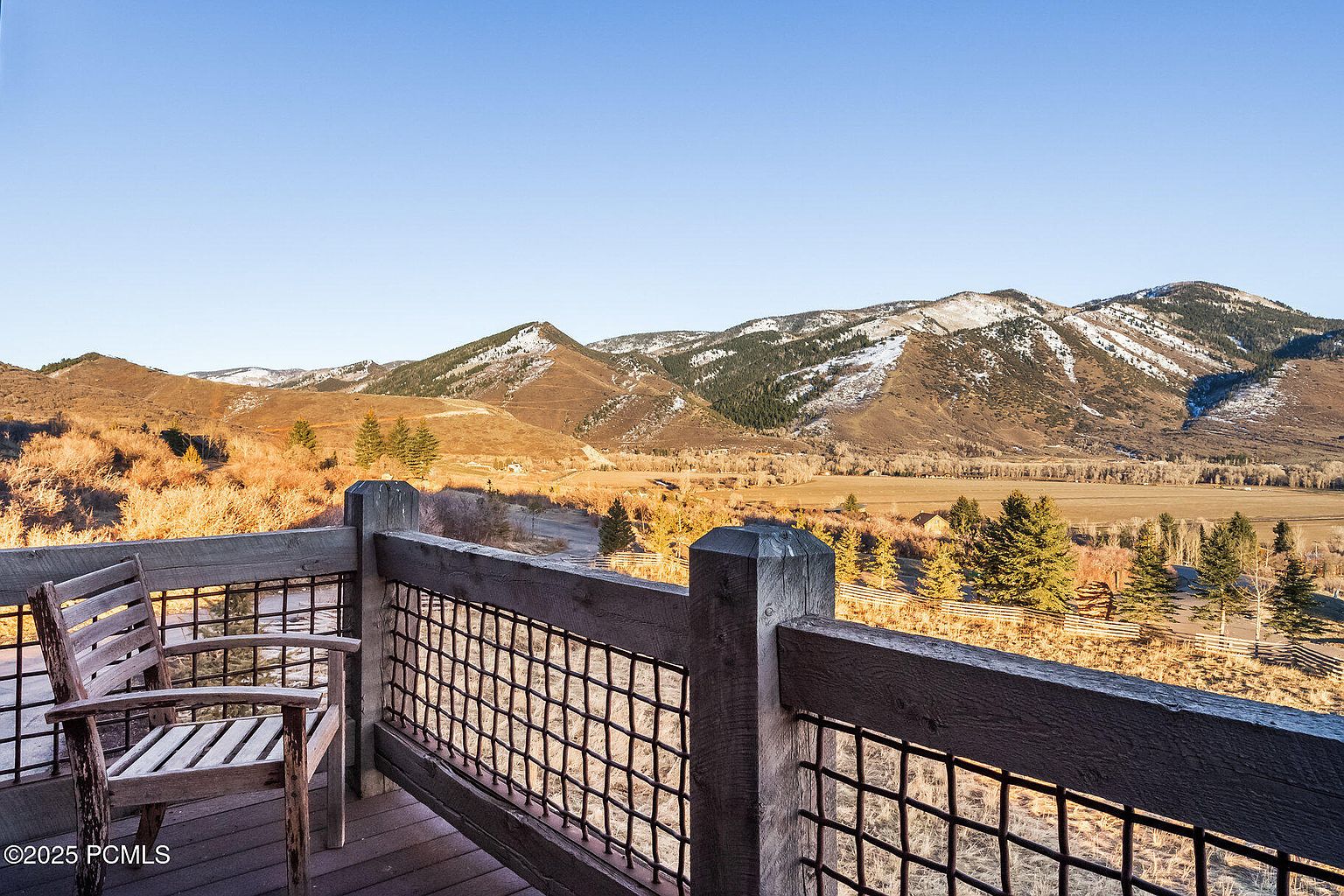 This image showcases a rustic wooden deck or balcony with a chair, offering a scenic view of rolling hills and mountains in the background. The deck features a wooden railing with a metal mesh design, adding to the rustic charm. The landscape includes a mix of trees and open fields, creating a serene and inviting atmosphere, perfect for enjoying the natural surroundings.