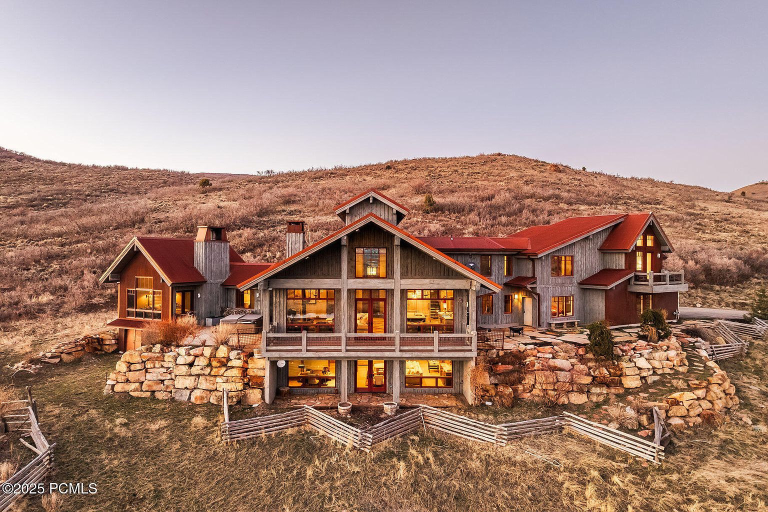 This aerial view showcases a sprawling, multi-level home nestled into a hillside. The residence features a red roof, gray wood siding, and numerous windows that offer ample natural light. Stone retaining walls and a rustic wooden fence add to the property's charm and blend it with the natural landscape.