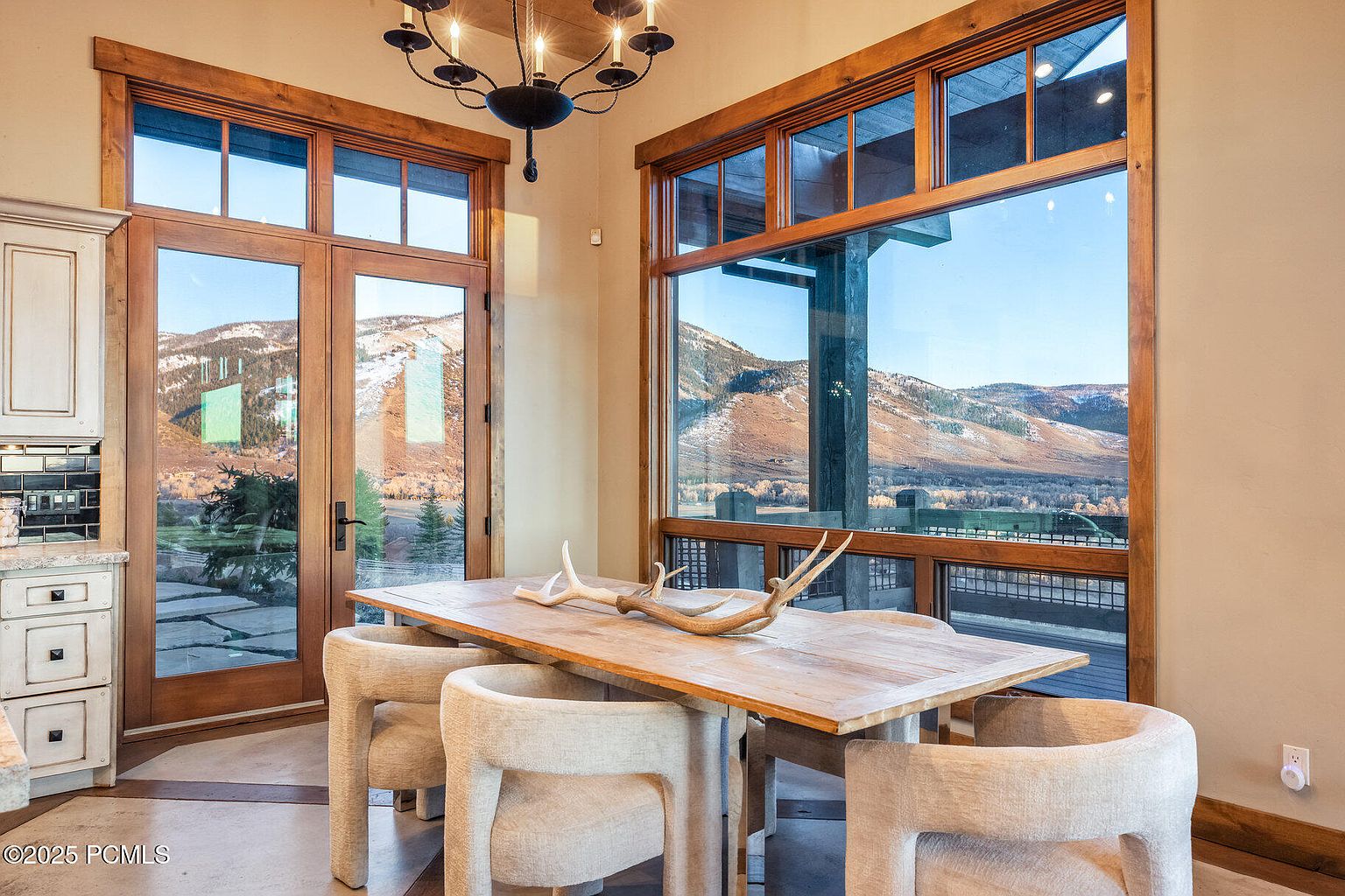 This dining room features a rustic wooden table with modern upholstered chairs, set against a backdrop of large windows offering scenic mountain views. A unique antler centerpiece adds a touch of natural elegance, while a chandelier provides warm lighting. The room combines natural elements with contemporary design, creating a welcoming and stylish space.