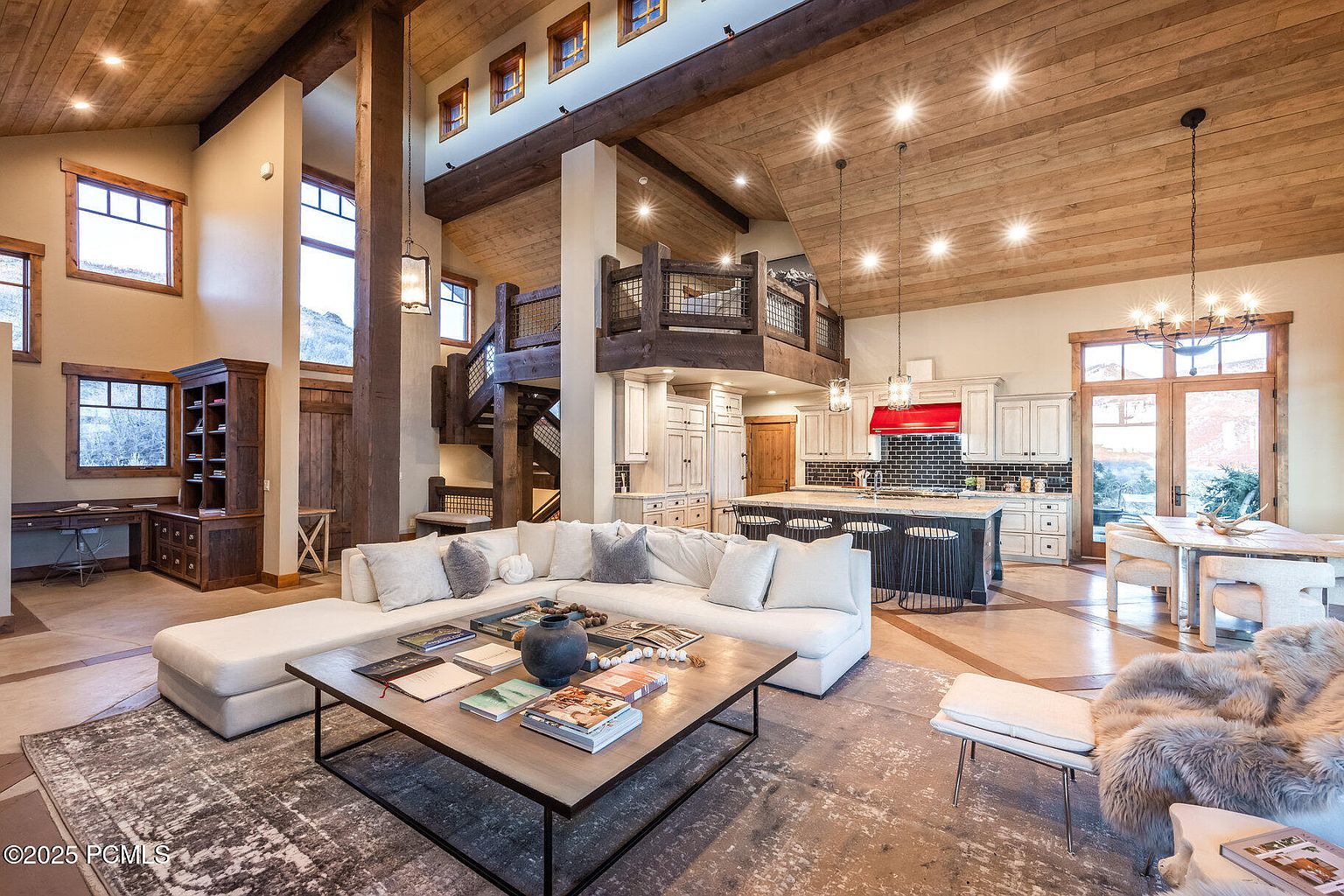 This is an interior shot of a luxurious living room that seamlessly blends into the kitchen and dining area. The living room features a large sectional sofa, a stylish coffee table with books, and a neutral-toned rug. The kitchen boasts modern appliances, a dark backsplash, and an island with bar seating, while the dining area is illuminated by a chandelier and natural light from the large windows. The open floor plan and high ceilings create a spacious and inviting atmosphere, perfect for entertaining.