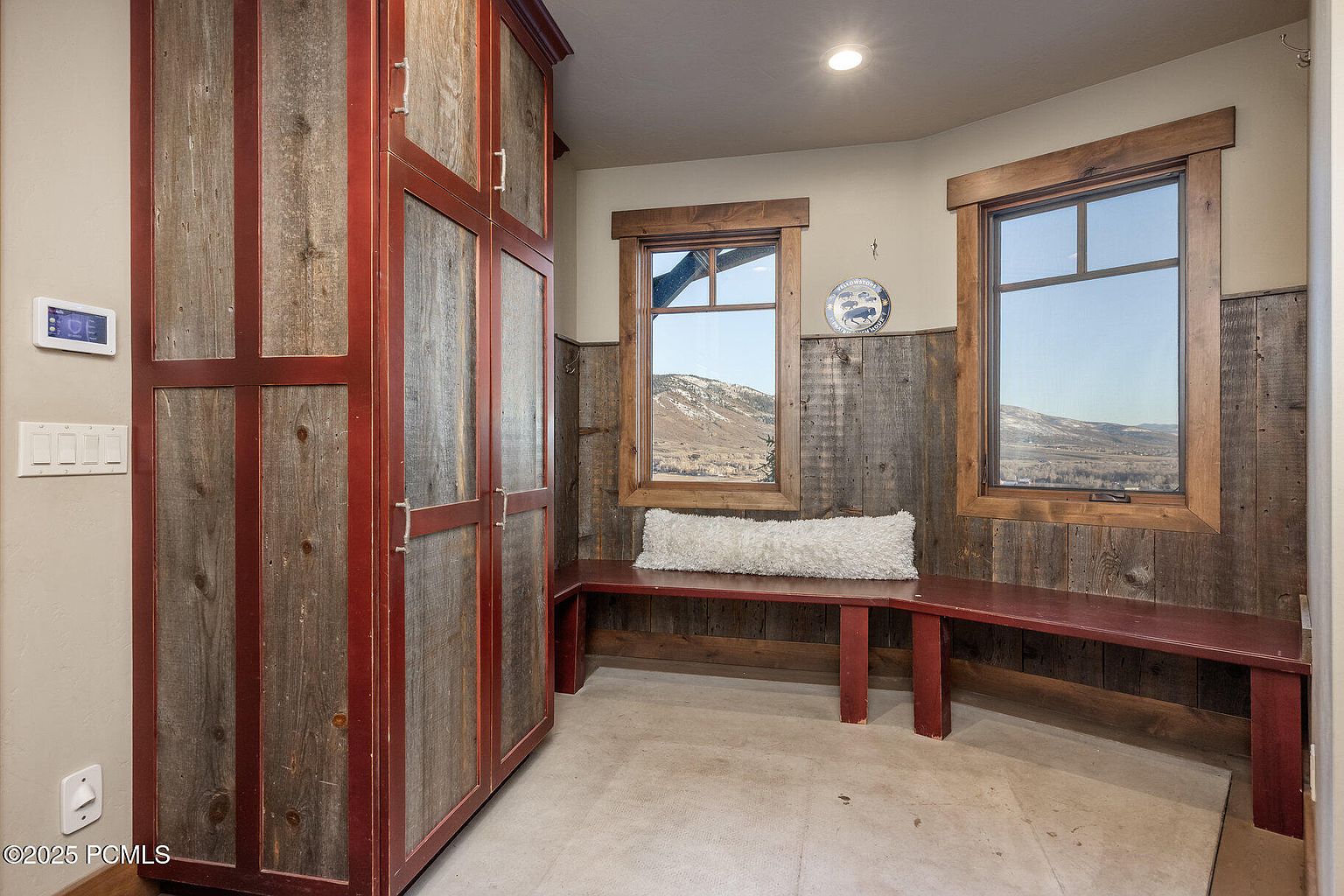This interior shot showcases a hallway or entryway featuring custom wooden cabinetry with a red trim, providing ample storage. A built-in bench sits beneath two windows, offering a view of the landscape outside. The walls are a neutral tone, complemented by wood paneling, creating a warm and inviting atmosphere.