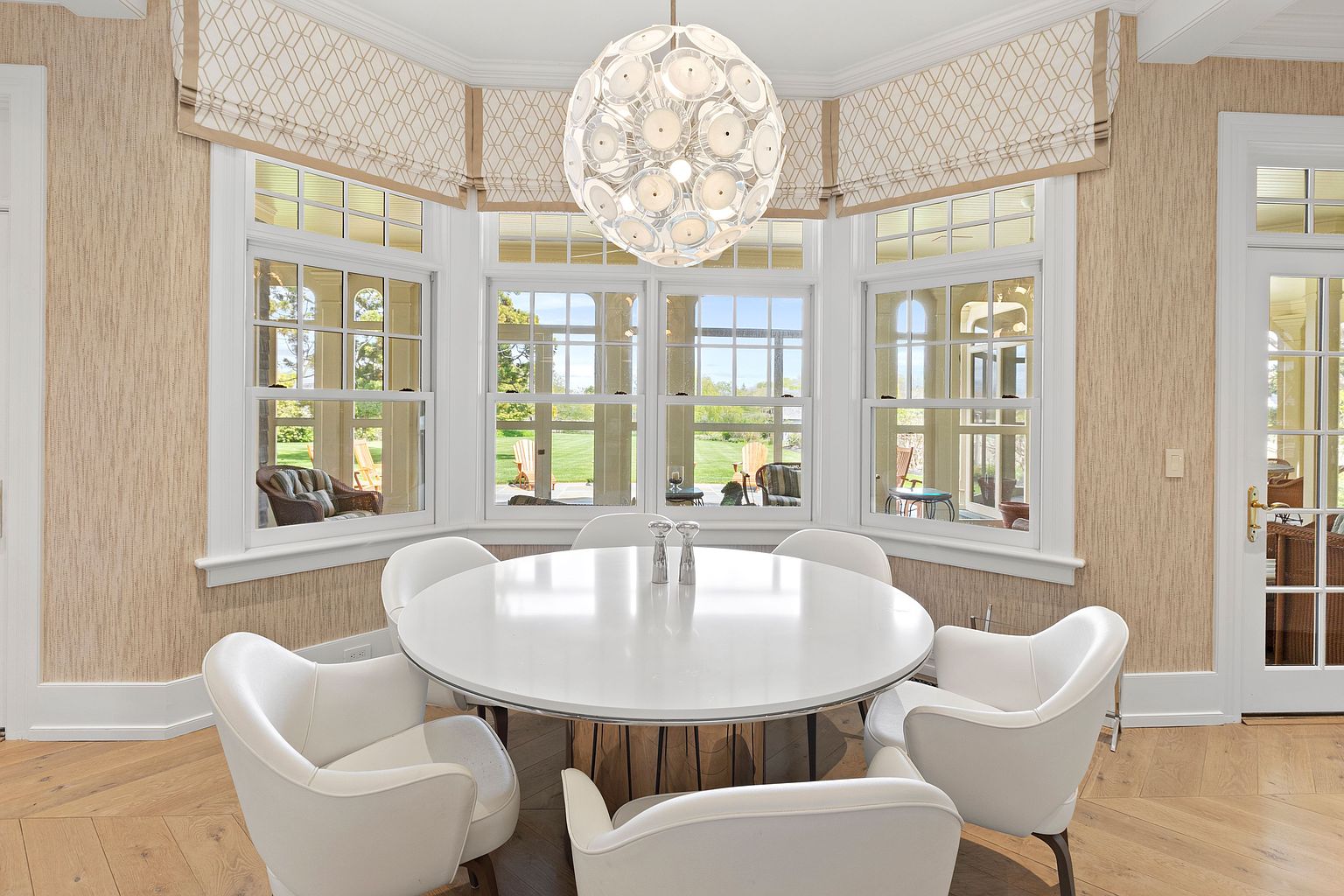 This bright dining area features a round white table surrounded by five modern, upholstered chairs with curved backs. A striking, spherical multi-light pendant fixture hangs above the table, while the room is framed by a bay of large windows that look out onto a lush green lawn. The space is finished with light wood flooring and textured, neutral-toned wallpaper, creating a sophisticated and airy atmosphere.