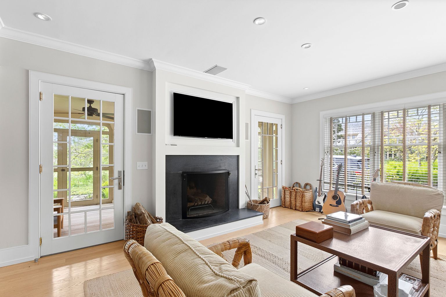 This inviting living room features a central fireplace with a dark stone surround and a mounted television above. The space is furnished with comfortable wicker-style seating and a wooden coffee table, all set upon a textured area rug. Large windows with blinds allow natural light to fill the room, which also includes glass-paned doors leading to an adjacent outdoor area.