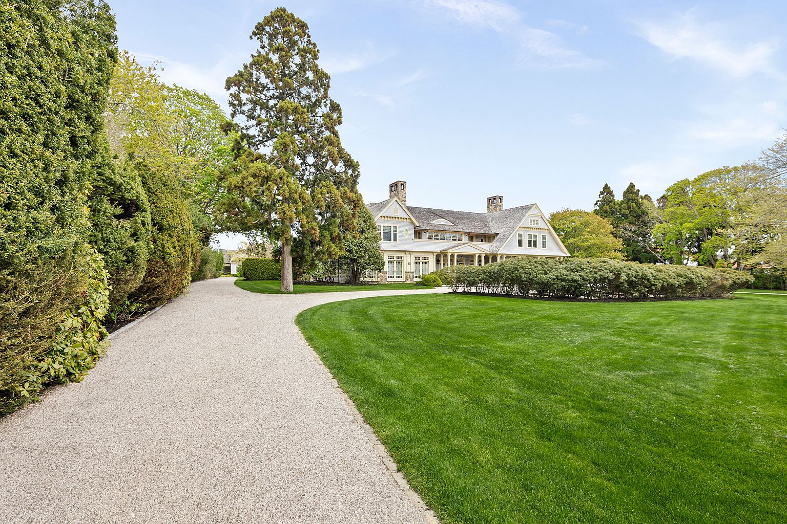 This expansive estate features a classic shingle-style residence set back from a winding gravel driveway. The property is surrounded by meticulously manicured lawns, mature trees, and dense, sculpted hedges that provide a sense of privacy and grandeur. The home's architecture includes multiple gables, stone accents, and a welcoming covered porch, creating a sophisticated and timeless curb appeal.
