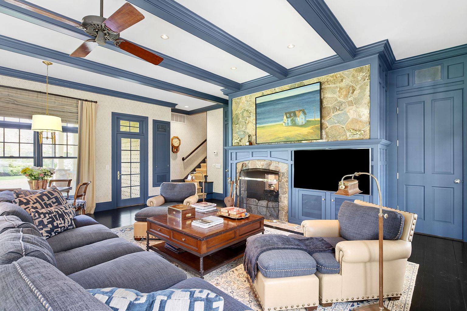 This inviting living room features a prominent stone-clad chimney breast housing a fireplace and a television, framed by deep blue built-in cabinetry and trim. The space is furnished with a large, plush sectional sofa and matching armchairs with ottomans, all arranged around a substantial wooden coffee table on a patterned area rug. A coffered ceiling with recessed lighting and a wooden ceiling fan adds architectural depth, while a doorway leads to a staircase in the background.