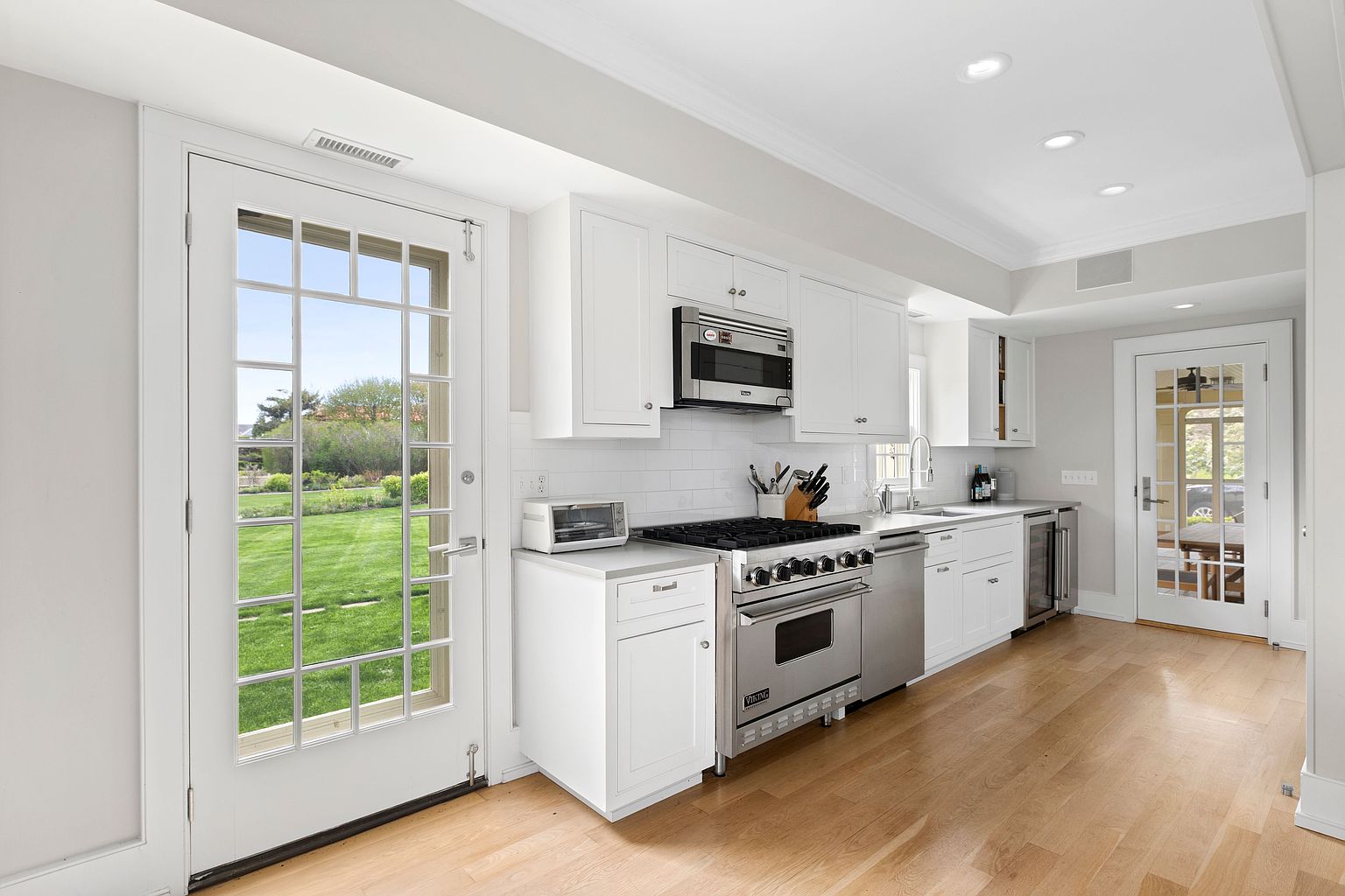 This bright, modern kitchen features crisp white cabinetry, a professional-grade stainless steel range, and light wood flooring. The space is illuminated by recessed lighting and natural light streaming through two glass-paned doors that provide views of the lush green yard. The clean aesthetic is complemented by a white subway tile backsplash and a functional layout that flows seamlessly toward the outdoor space.