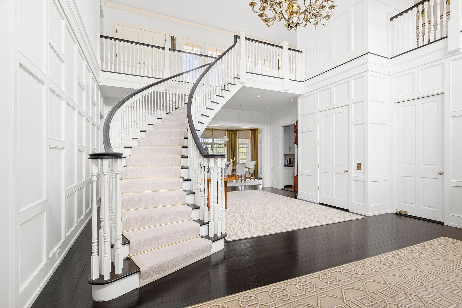 This grand foyer features a dramatic curved staircase with a dark wood handrail and white balusters, serving as the focal point of the space. The area is finished with extensive white wall paneling, dark hardwood flooring, and a large, intricate chandelier hanging from the high ceiling. A view through the open archway reveals a glimpse of a dining area, creating an inviting and elegant transition between rooms.