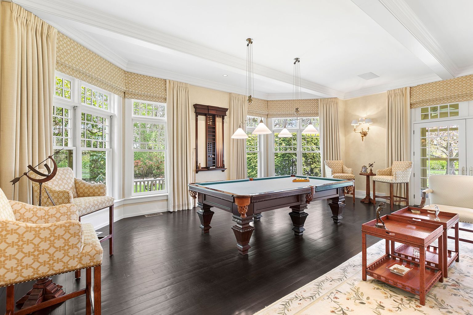 This elegant game room features a central billiard table set against a backdrop of large, multi-pane windows that offer views of the surrounding greenery. The space is furnished with patterned armchairs, a tiered wooden side table, and a wall-mounted cue rack, all set upon dark hardwood flooring. The room is illuminated by a combination of natural light and classic pendant lighting, creating a sophisticated and inviting atmosphere for recreation.