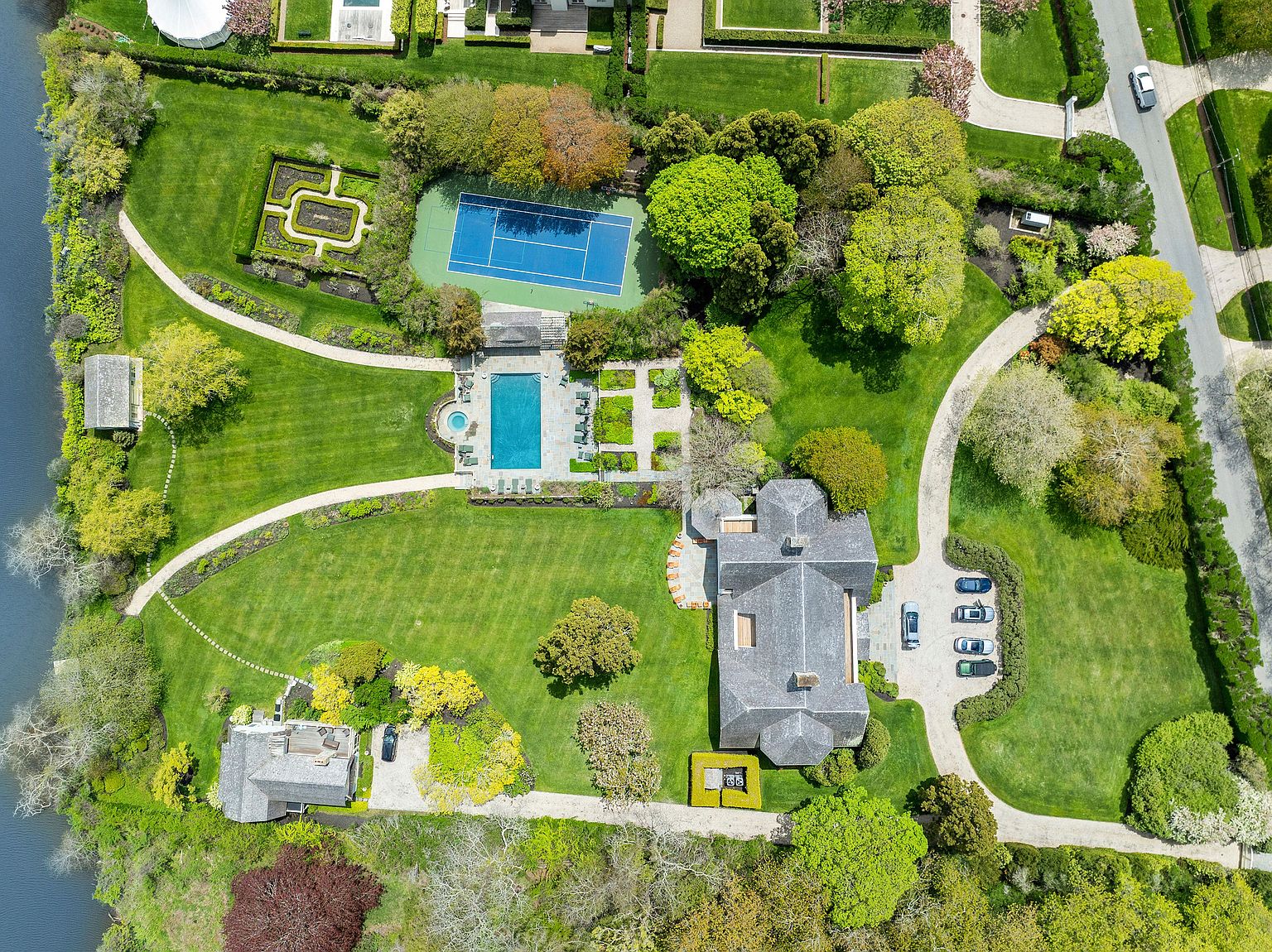 This expansive aerial view captures a sprawling, lush estate situated along a waterfront. The property features a large main residence, a separate guest house, a private tennis court, and a rectangular swimming pool surrounded by manicured lawns and mature trees. The winding driveway and structured garden paths create a sense of privacy and refined elegance, showcasing a classic, high-end residential landscape.