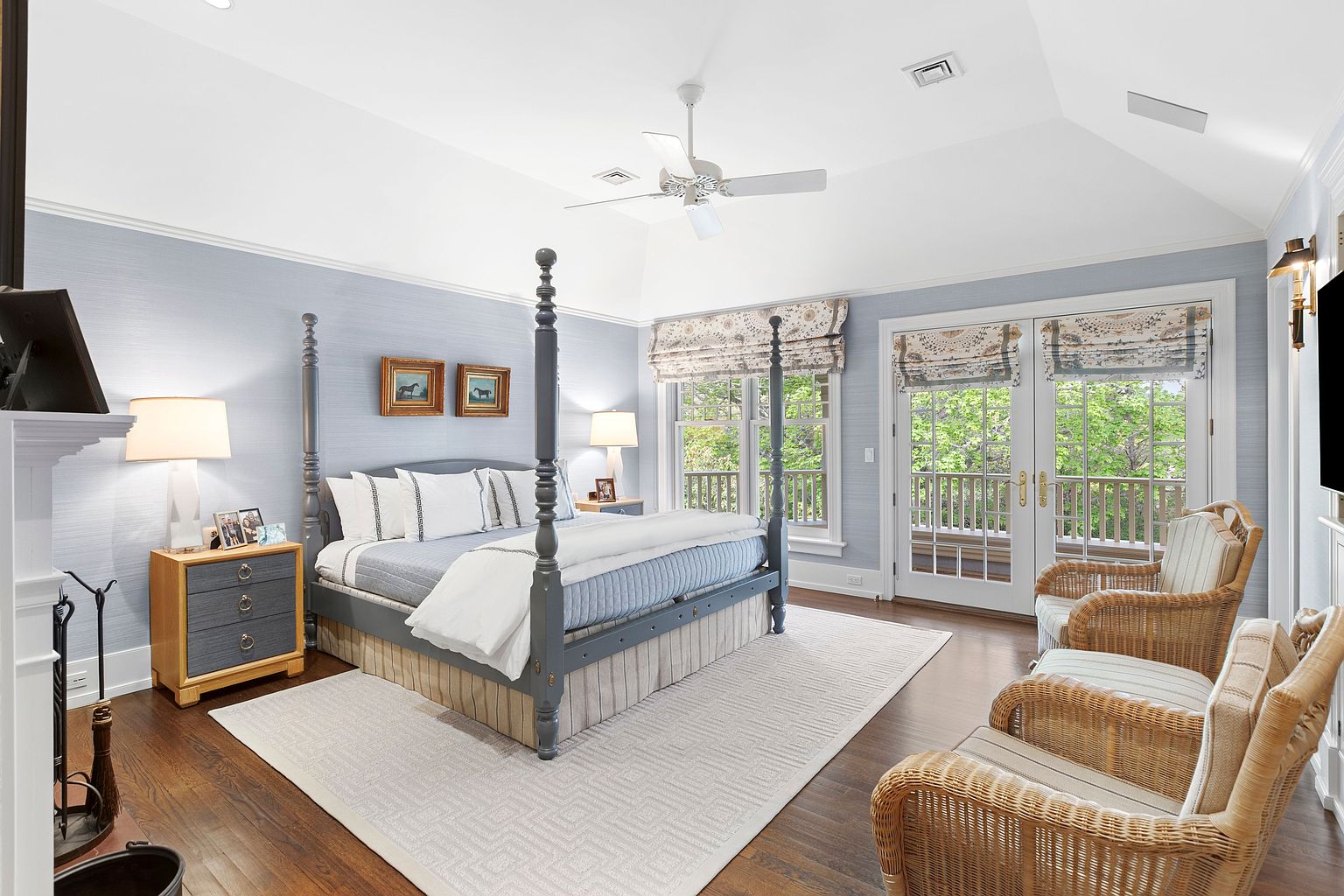 This spacious primary bedroom features a prominent four-poster bed as the central focal point, set against light blue textured wallpaper. The room includes a cozy seating area with two wicker armchairs, a fireplace on the left, and glass doors that open to an outdoor balcony. The vaulted ceiling and dark wood flooring contribute to an elegant, airy atmosphere.