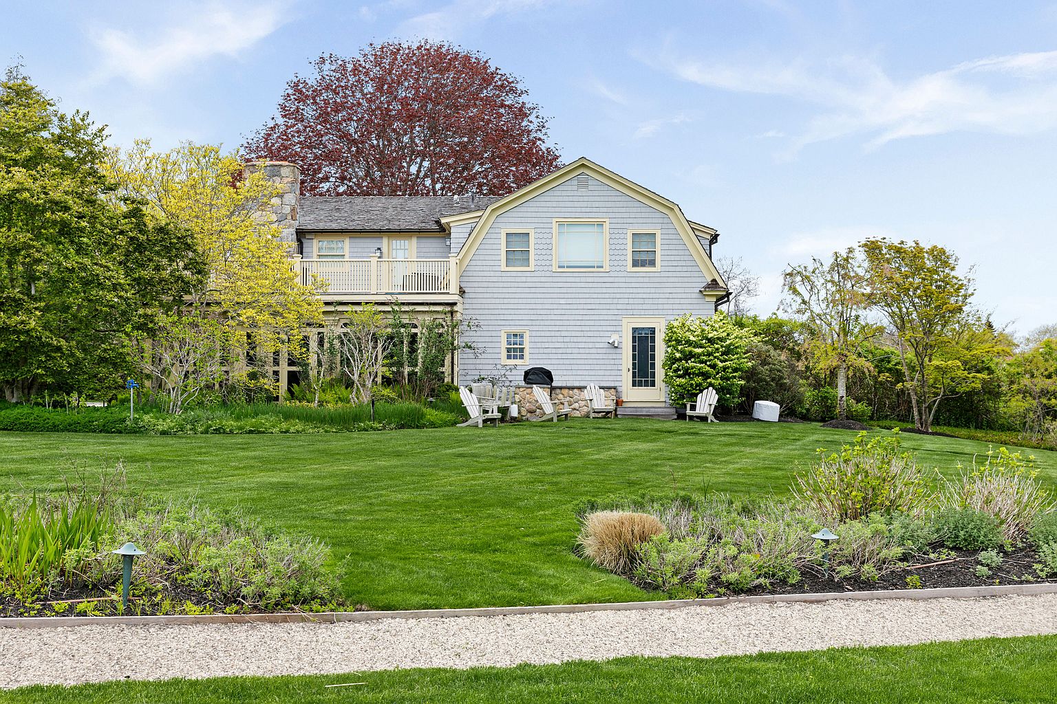 This rear view of a charming shingle-style home showcases a gambrel roofline and a stone-clad chimney breast. The property features a spacious, well-manicured lawn, a gravel path in the foreground, and a second-story balcony overlooking the garden. Several Adirondack chairs are arranged on the lawn, creating a peaceful outdoor seating area against the backdrop of mature trees and lush landscaping.