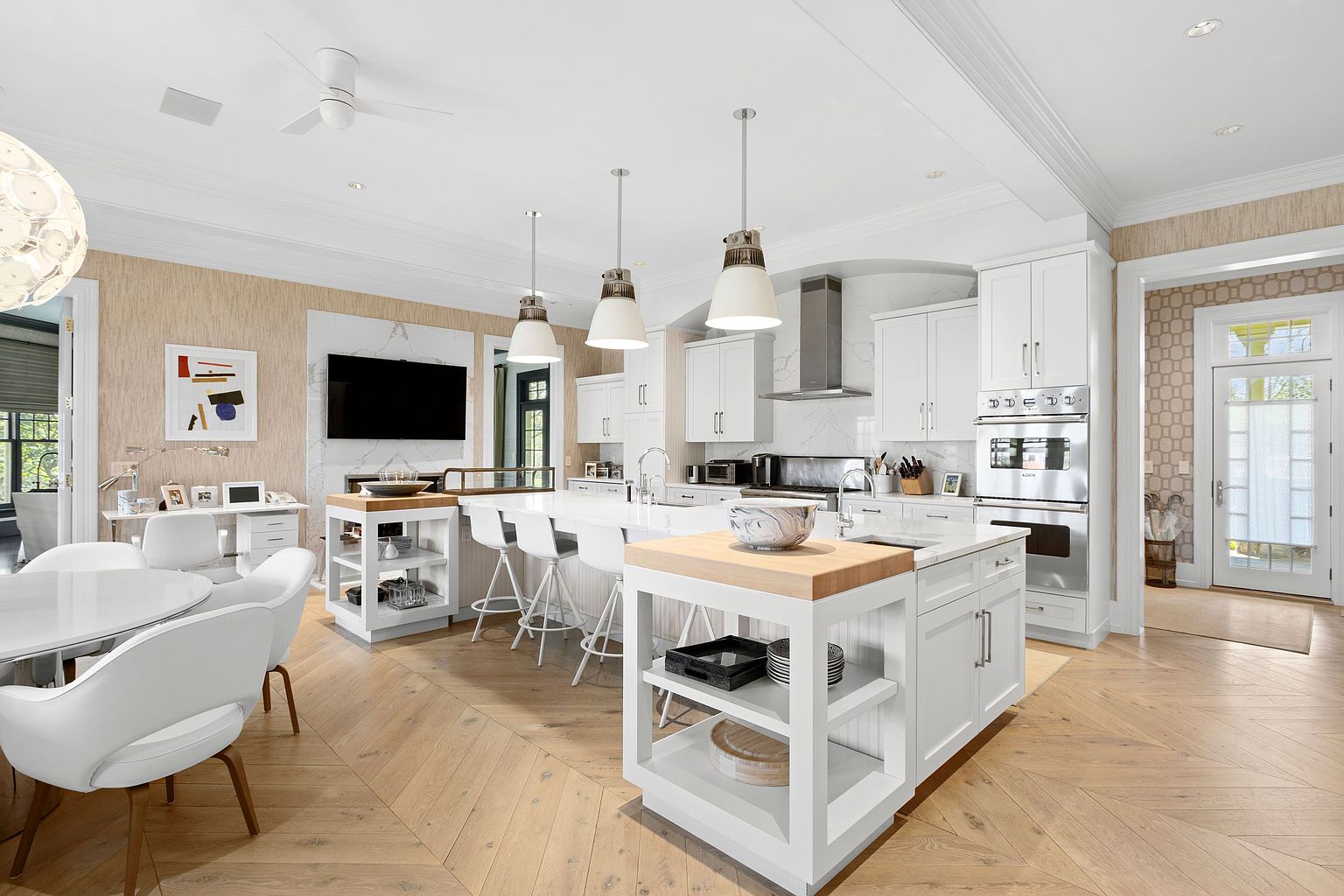 This bright and airy kitchen features a large central island with a light wood butcher block top and white cabinetry, complemented by a secondary island with bar seating. The space is anchored by light-toned wood flooring arranged in a chevron pattern and includes a dedicated desk area, a wall-mounted television above a stone-clad chimney breast, and a dining nook. Three pendant lights hang over the main island, creating a cohesive and inviting atmosphere that flows into an adjacent entryway.