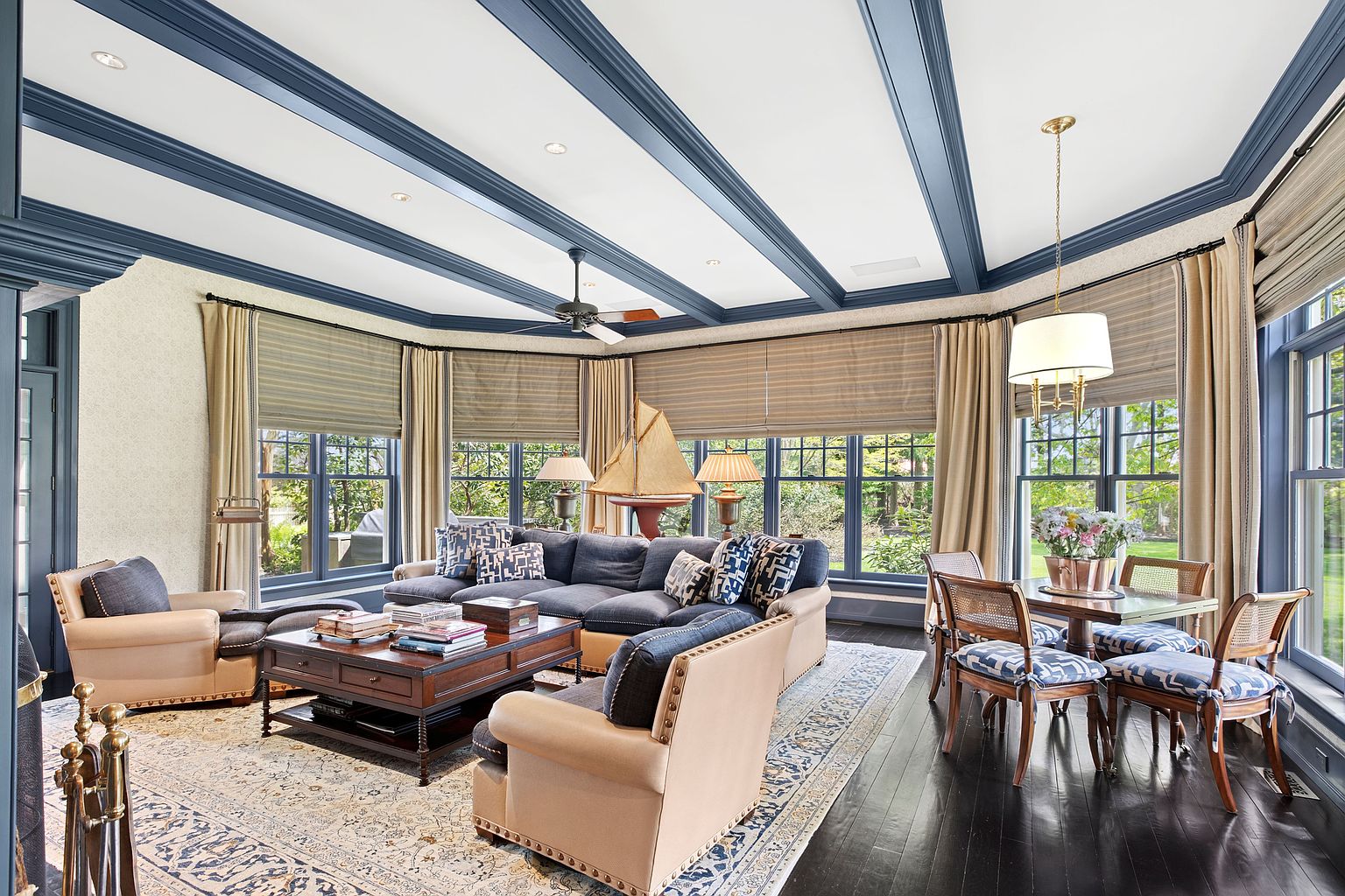 This elegant living room features a spacious layout with a large, dark-toned sectional sofa and matching armchairs arranged around a substantial wooden coffee table. The room is defined by its striking blue-painted coffered ceiling and expansive windows that provide abundant natural light and views of the surrounding greenery. A dining area with a round table and cane-back chairs sits to the side, while a decorative model sailboat serves as a focal point on the sofa table, creating a sophisticated and inviting atmosphere.