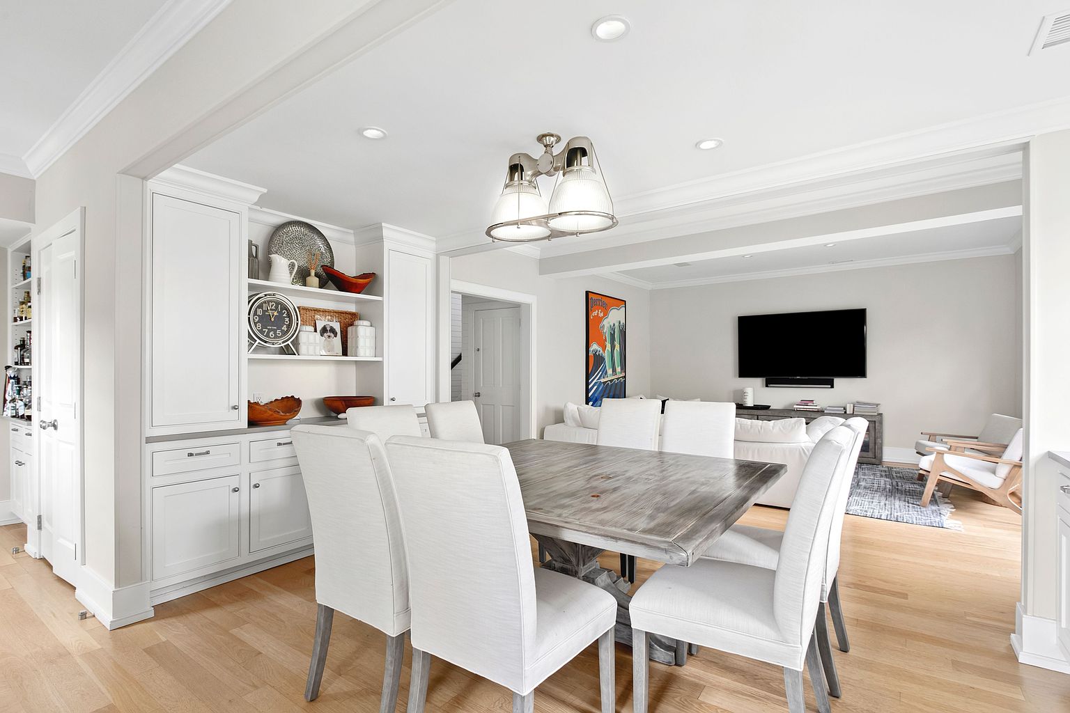 This bright and open dining area features a rustic wood table surrounded by six upholstered chairs, positioned beneath a modern three-light fixture. To the left, a built-in white cabinetry unit provides ample storage and display space, while the room flows seamlessly into a living area with a large television and comfortable seating. The space is finished with light wood flooring and neutral walls, creating a cohesive and inviting atmosphere.