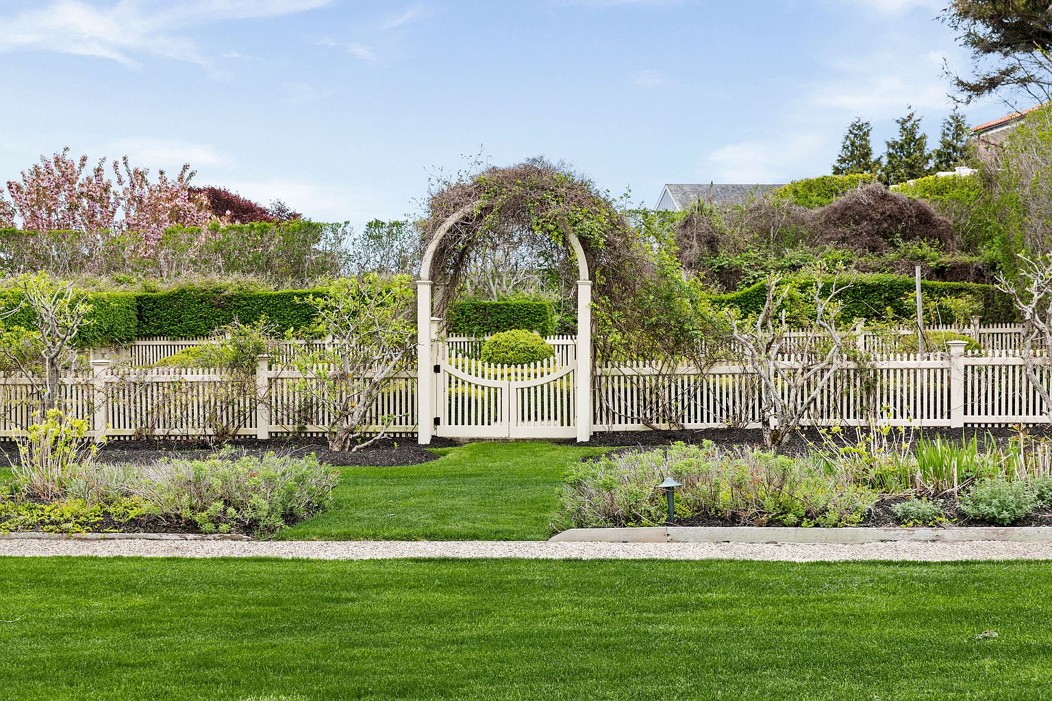 This beautifully manicured garden features a central white picket fence with an arched gate covered in climbing vines. The foreground showcases a lush green lawn and a gravel path, while the background is framed by mature hedges and flowering trees. The scene offers a serene and classic aesthetic, perfect for a peaceful outdoor retreat.