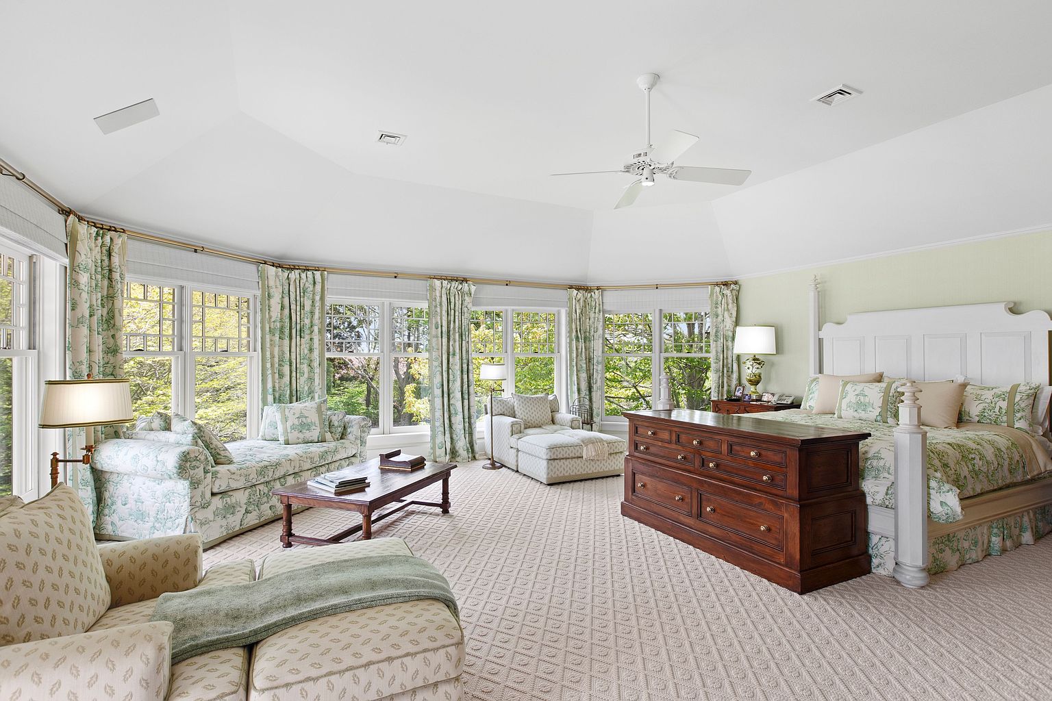 This spacious primary bedroom features a vaulted ceiling with a central white ceiling fan and a large, curved window wall that floods the room with natural light. The space is furnished with a large bed, a dark wood storage chest at the foot of the bed, a patterned chaise lounge, and a cozy seating area with a coffee table. The room is decorated in a classic, soft-toned aesthetic with patterned textiles and light-colored carpeting, creating a serene and inviting atmosphere.
