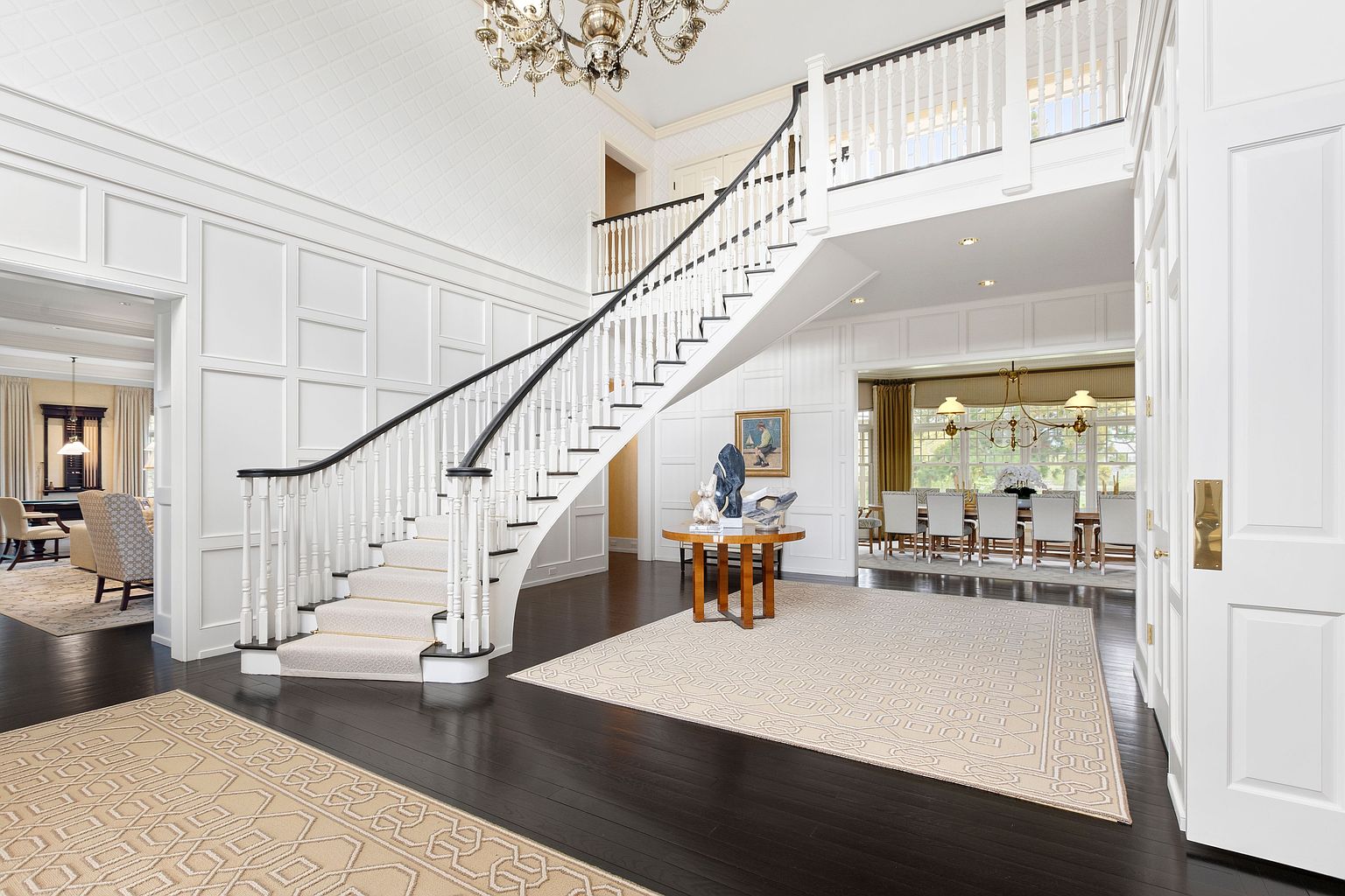This grand foyer features a sweeping staircase with a dark wood handrail and white balusters, serving as the focal point of the space. The area is finished with crisp white wall paneling, dark hardwood flooring, and a large, intricate chandelier hanging from the high ceiling. A view into the adjacent dining room and a glimpse of a living area create an open, airy, and sophisticated atmosphere.