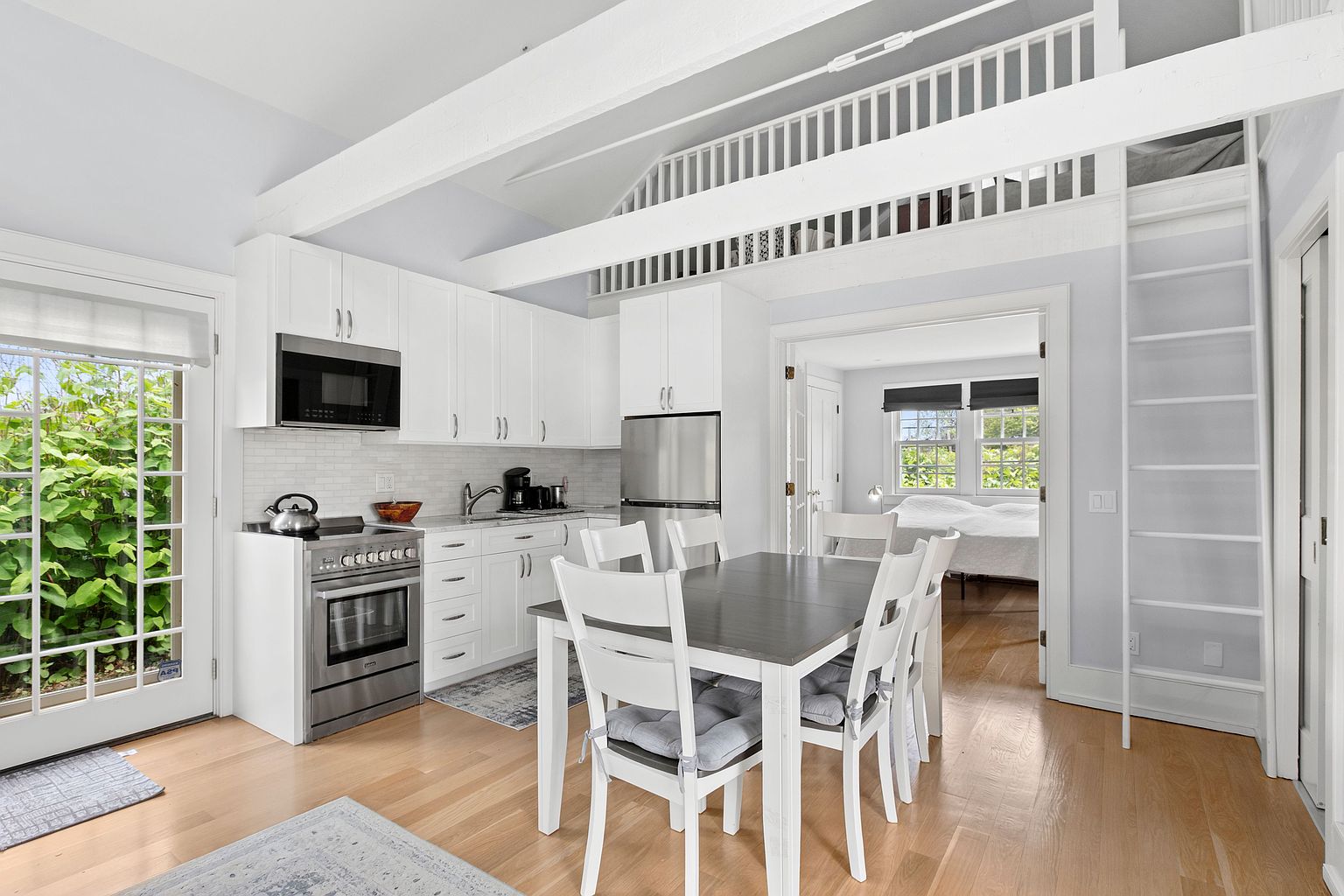 This bright, open-concept kitchen features white cabinetry, stainless steel appliances, and light wood flooring. A dining table with white chairs sits centrally, while a ladder leads to a lofted space above, creating a cozy and functional living area. The room is illuminated by natural light streaming through a glass-paned door and a doorway leading into an adjacent bedroom.