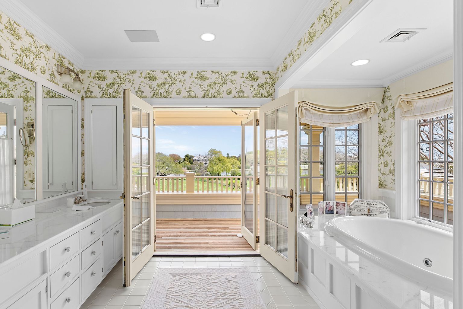This elegant primary bathroom features a double vanity with white cabinetry and a light-toned stone countertop, complemented by patterned wallpaper. A deep soaking tub is positioned beneath large windows, while French doors open directly onto a private wooden balcony overlooking a scenic waterfront view. The space is bright and airy, blending classic design elements with a seamless transition to the outdoors.