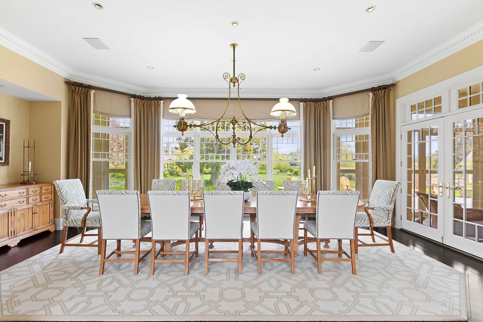 This elegant dining room features a long wooden table surrounded by upholstered chairs with a subtle patterned fabric. A large, ornate brass chandelier hangs centrally above the table, while a geometric-patterned area rug anchors the space. The room is framed by a curved wall of large windows draped in neutral curtains, offering a bright and airy view of the lush green landscape outside.