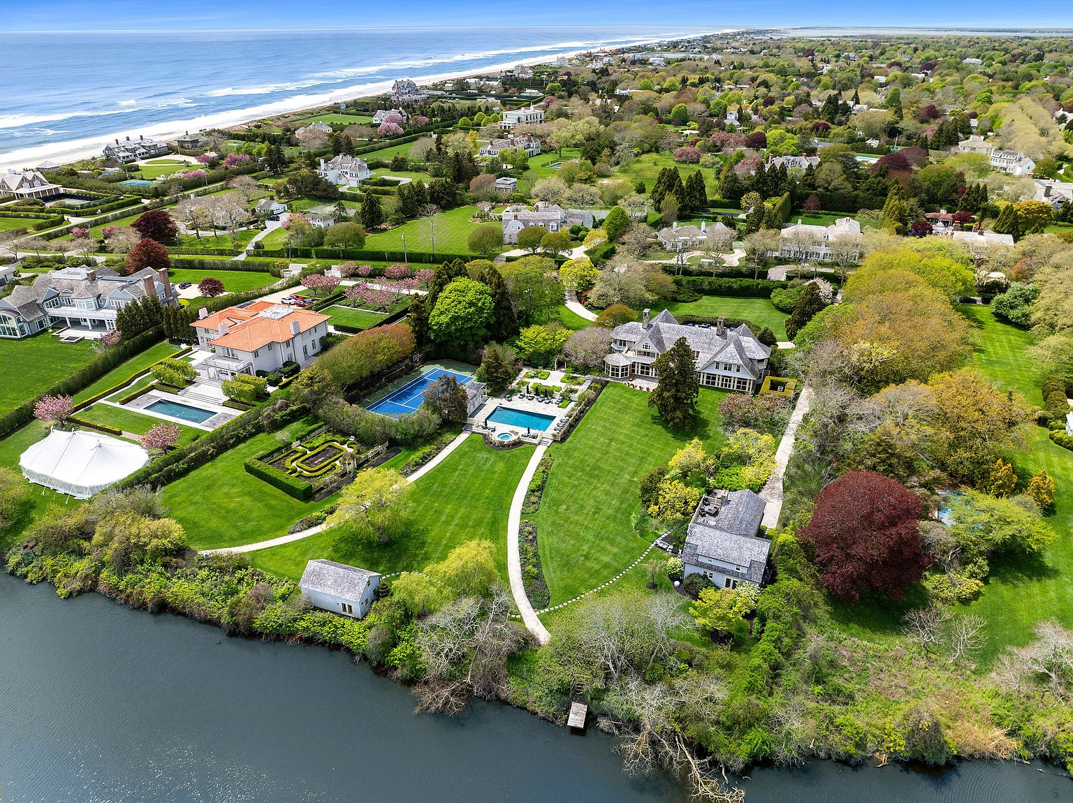 This expansive aerial view captures a luxurious coastal estate featuring multiple large residences, manicured lawns, and private swimming pools. The property includes a tennis court, formal garden beds, and a winding path leading toward a serene waterfront edge. The scene is set against a backdrop of lush greenery and the distant Atlantic coastline, emphasizing the prestigious and secluded nature of the location.