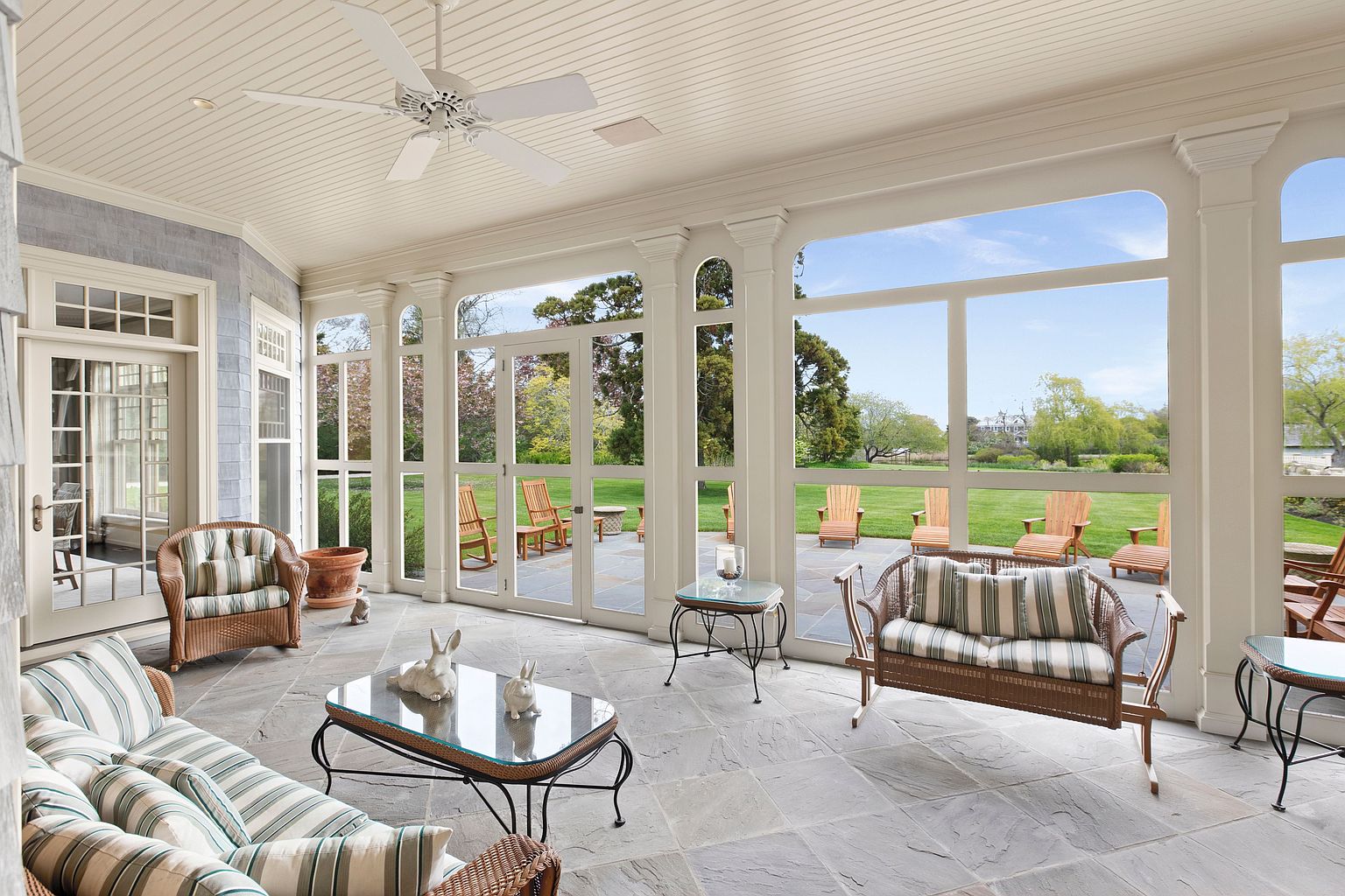 This bright and airy sunroom features a stone-tiled floor and a white beadboard ceiling with a central ceiling fan. The space is furnished with comfortable wicker seating featuring striped cushions and a glass-topped coffee table, while large windows provide an expansive view of the lush green lawn and patio outside. The room offers a serene, transitionary space that seamlessly connects the indoor living area with the outdoor landscape.