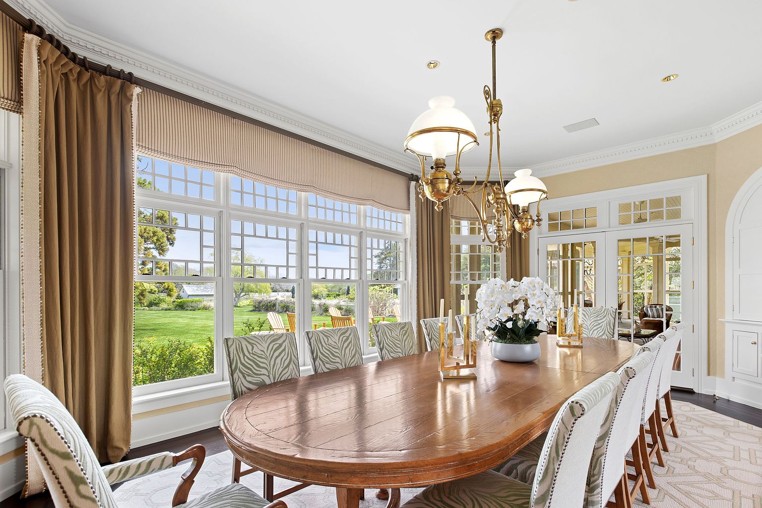 This elegant dining room features a large, oval wooden table surrounded by chairs with patterned upholstery, set beneath a classic brass chandelier. A wall of large windows with striped valances and brown curtains provides abundant natural light and views of the lush green lawn outside. The space is finished with crown molding, a patterned area rug, and a set of glass-paned French doors leading to an adjacent room.